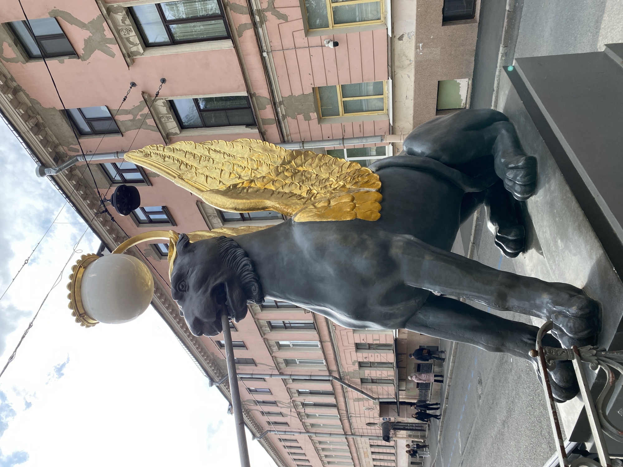 A low-angle, close-up shot depicts the Gryphon statue on Universitetskaya Embankment in Saint Petersburg, Russia. The gryphon, a mythical creature with the body of a lion and wings of an eagle, is prominently featured. Its body is a dark color, likely bronze or iron, with golden wings. The gryphon also functions as a light post, with a large, spherical lamp attached to its head.
In the background, a building with multiple windows is visible, its facade painted in shades of pink and beige. Along the street, several people are walking, though they are seen from a distance and their features are not clearly defined.
The sky is partially visible, with a mix of clouds and blue sky. The photograph's angle gives a sense of the gryphon's elevation and prominence in the urban landscape.