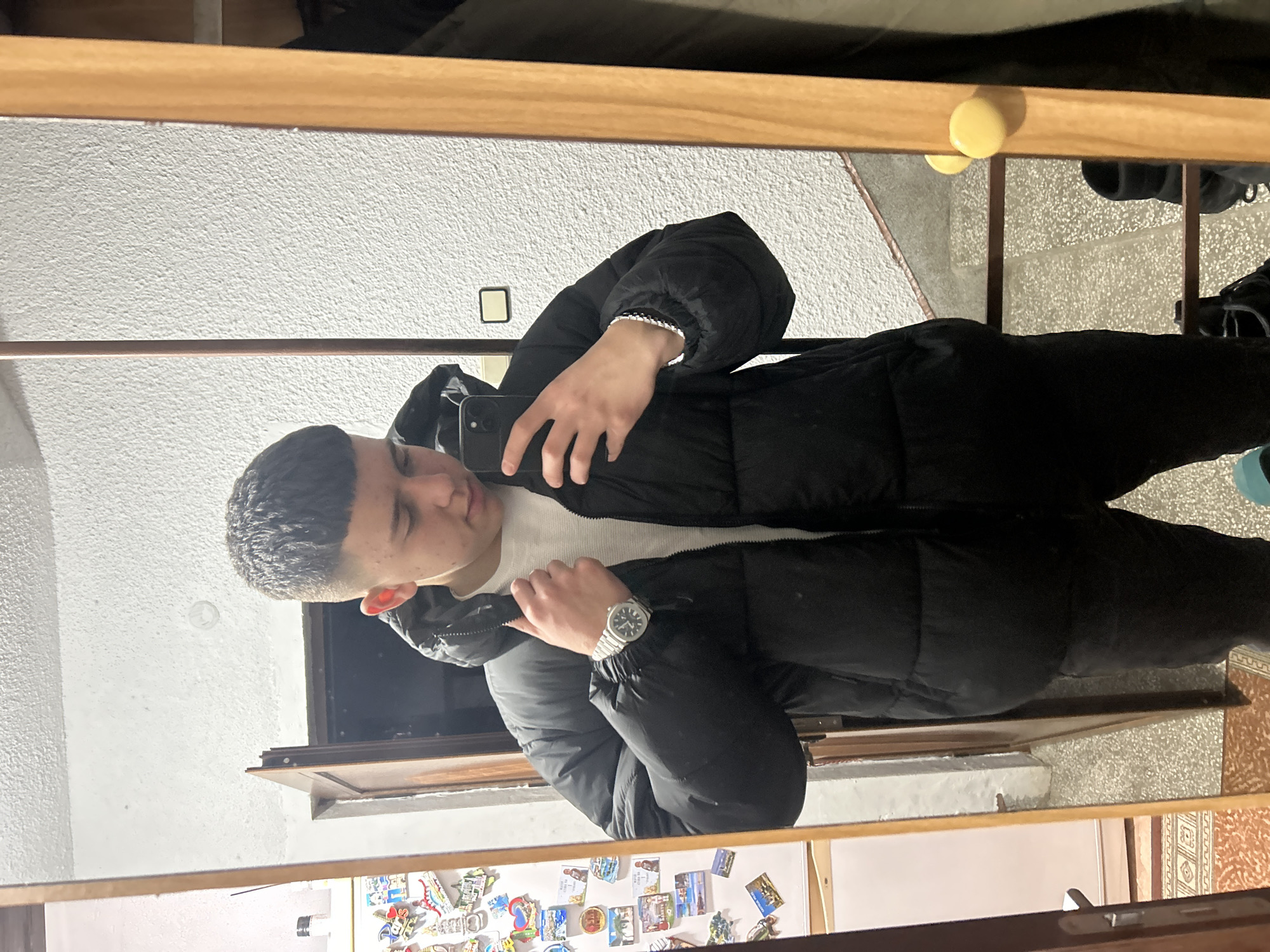 A young man stands in front of a mirror, likely taking a selfie. He is wearing a black puffer jacket over a white shirt and a silver wristwatch. The reflection shows a room with a white wall behind him, and the bottom of the mirror is adorned with magnets, possibly souvenirs. Based on the available details, pinpointing the exact location in Zlatograd, Bulgaria, is impossible.