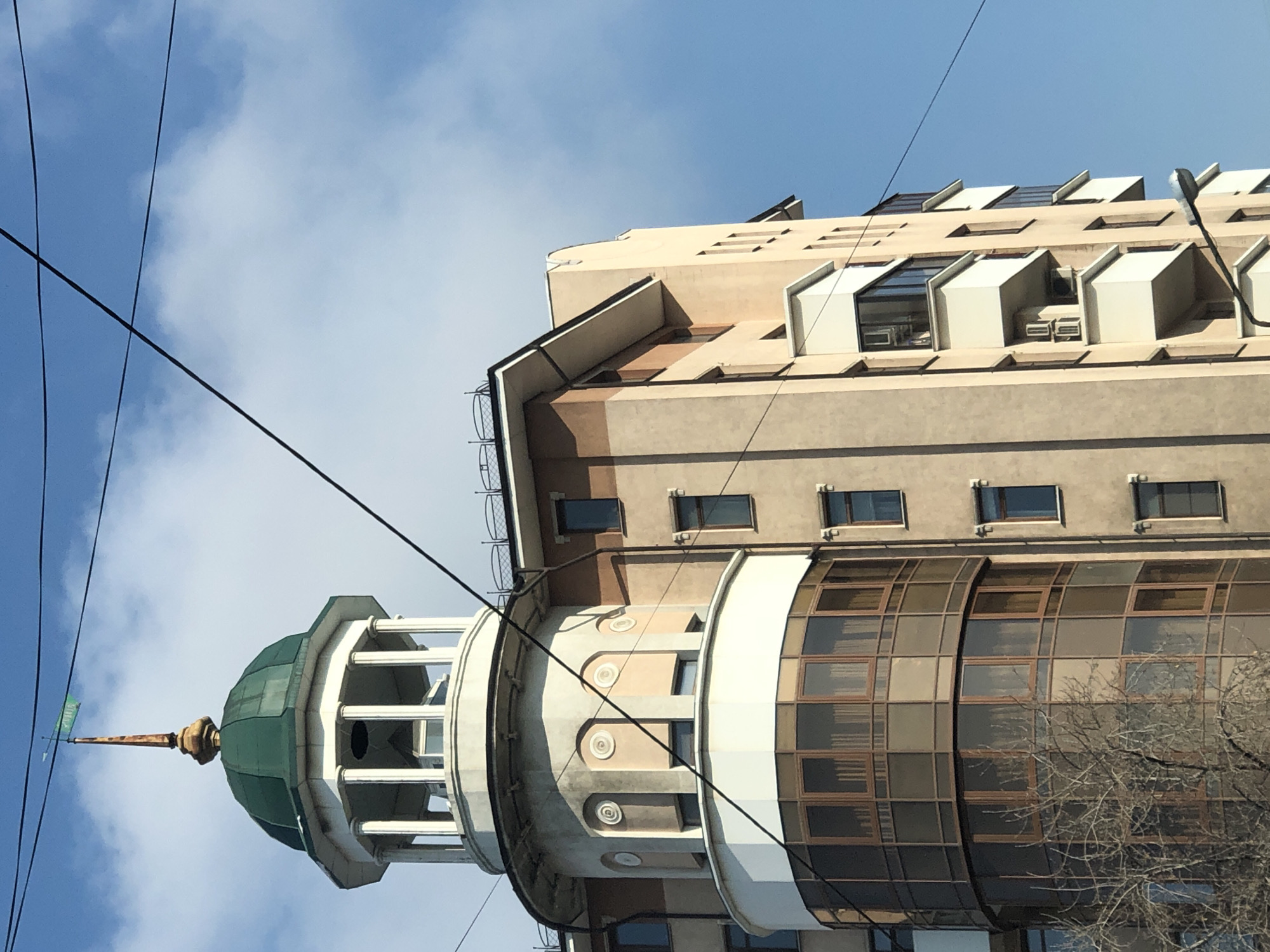 This image presents an upward view of a distinctive multi-story building in Krasnoyarsk, Russia, set against a blue sky with scattered white clouds. The building showcases varied architectural elements. The lower section features a prominent, curved facade composed of numerous brown-tinted glass panes within a grid framework. Above this, a rounded part of the structure is adorned with decorative circular motifs and small rectangular windows, topped by a white band.
Crowning the building is a green, multi-faceted dome, which rests on a white cupola supported by columns. From the dome extends a slender spire, gold-colored at its base, tapering to a point and topped with a small green weather vane. The uppermost floors are rectangular, constructed from a light tan material, with a roofline featuring multiple skylights or dormer windows, some enclosed within white, angular box-like structures, possibly serving as sunshades or unique architectural accents.
Several black power lines crisscross the frame, partially obscuring portions of the building and the sky. Bare tree branches are visible in the lower right, extending over the glass facade. The scene depicts a clear and sunny daytime. On the green weather vane, a faint, illegible symbol or text can be discerned.