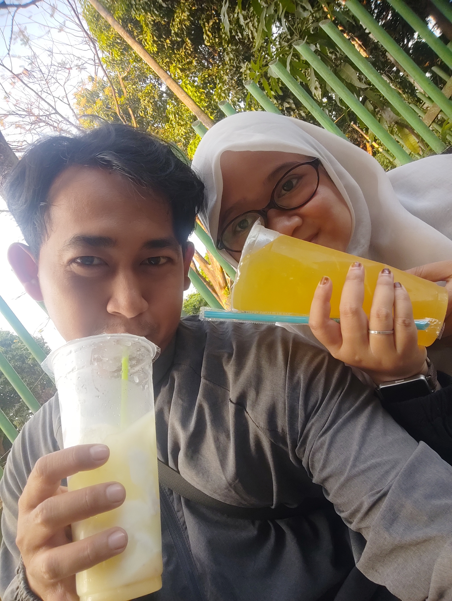 Two individuals, a man and a woman wearing a hijab, are posing for a selfie outdoors, likely in Cirebon, Indonesia. The man in the foreground is holding a plastic cup with a yellow drink and a straw, bringing it to his lips as if to drink. He is wearing a dark grey long-sleeved shirt. The woman, positioned behind and to the right of the man, is also holding a clear plastic bottle filled with a yellow liquid and a blue straw. She is wearing glasses and a white hijab. Both appear to be in good spirits. The background reveals a lush green environment with trees and a green fence, suggesting a park or outdoor recreational area. The lighting indicates it is daytime, possibly late afternoon given the angle of the sun.