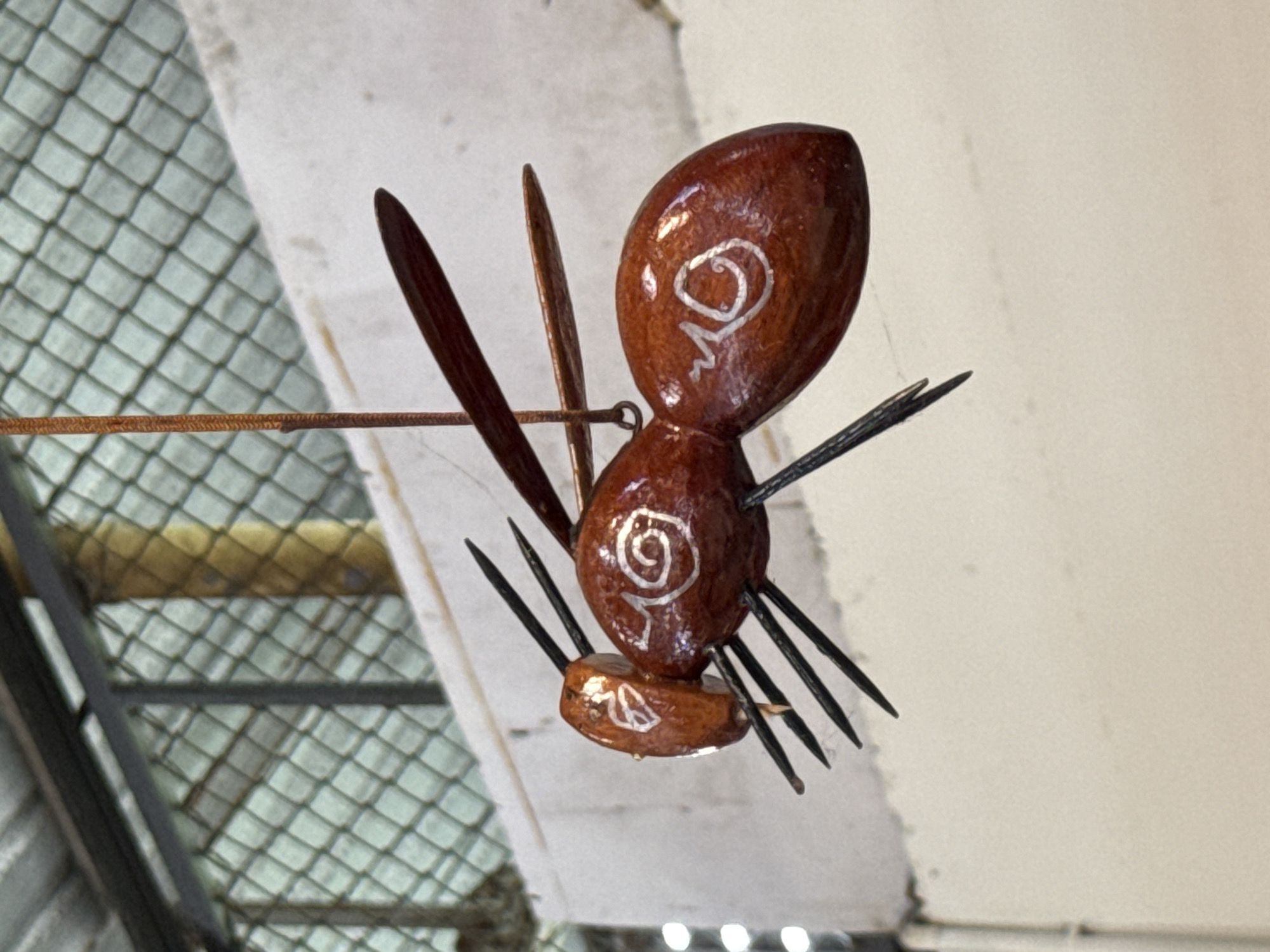 A wooden figurine of an insect, possibly a bee or wasp, is suspended by a thin string against a neutral background. The figurine is predominantly a reddish-brown color with white swirl patterns on its abdomen and head. It has six black, pointed legs and two pairs of reddish-brown wings. The background appears to be an indoor setting, with a corrugated metal roof and a chain-link fence visible in the upper portion of the image. The overall impression is of a decorative item displayed in a workshop or market environment. The exact location in Baan Bo Phut, Thailand, is not discernible from the visual elements of the image alone.