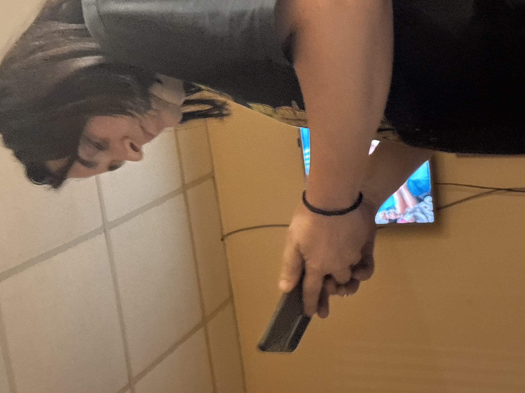 A person, seen from the side, is looking down at a smartphone held in their hands. They are wearing a dark t-shirt and a black bracelet on their right wrist. In the background, a television screen displays an image of a person with blonde hair. The scene appears to be indoors, with a textured ceiling and yellow walls. There is a cable running down the wall from the ceiling to the television. The location is specified as Tervel, Bulgaria.