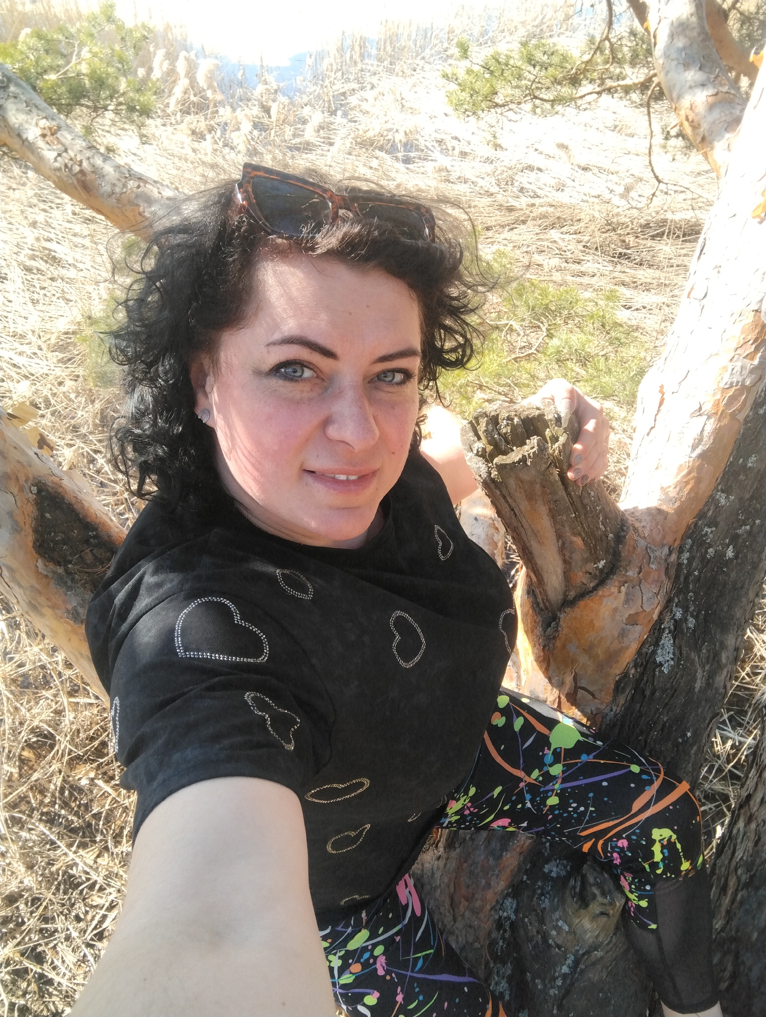 A woman is posing for a selfie outdoors. She has dark, curly hair and is wearing sunglasses on her head. She is wearing a black shirt with heart-shaped patterns and colorful leggings. She is sitting in a tree with light brown bark. The background consists of dry grass and some green vegetation. The location is Votkinsk, Russia.
