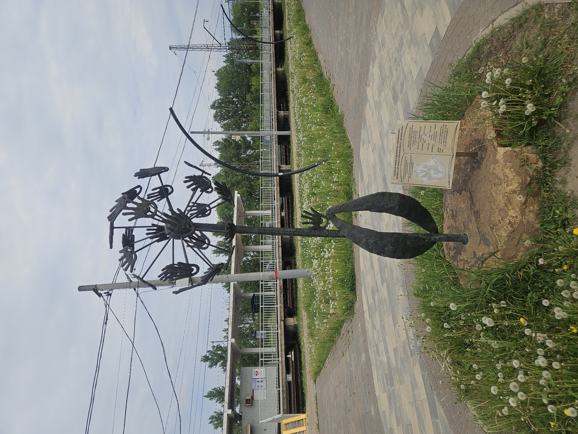 The image shows a metal sculpture in Речицы, Russia, near a railway line. The sculpture is of a dandelion seed head, but instead of seeds, it has hands at the ends of the spokes. The sculpture rests on a rock in a patch of grass dotted with actual dandelions. Beside the sculpture is a small plaque with text in Russian and English, describing the sculpture as a "Symbol of Peace and Kindness." In the background is a railway platform with overhead power lines. The walkway next to the train is made of light gray rectangular pavers. The sky is overcast.