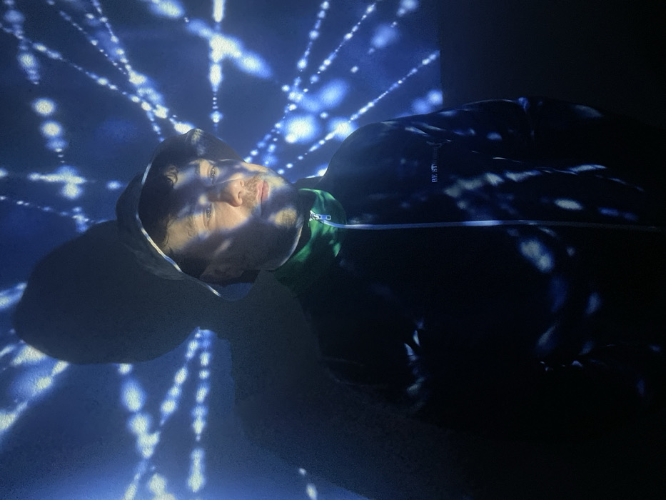 A man lies on his back, his head tilted slightly. He wears a hooded sweatshirt with a white drawstring and green accents. The background and his body are illuminated with a pattern of blue and white lights resembling connected dots and lines. The lighting casts shadows across his face and clothing, creating contrast and a sense of depth. The image appears to be taken at an angle.
