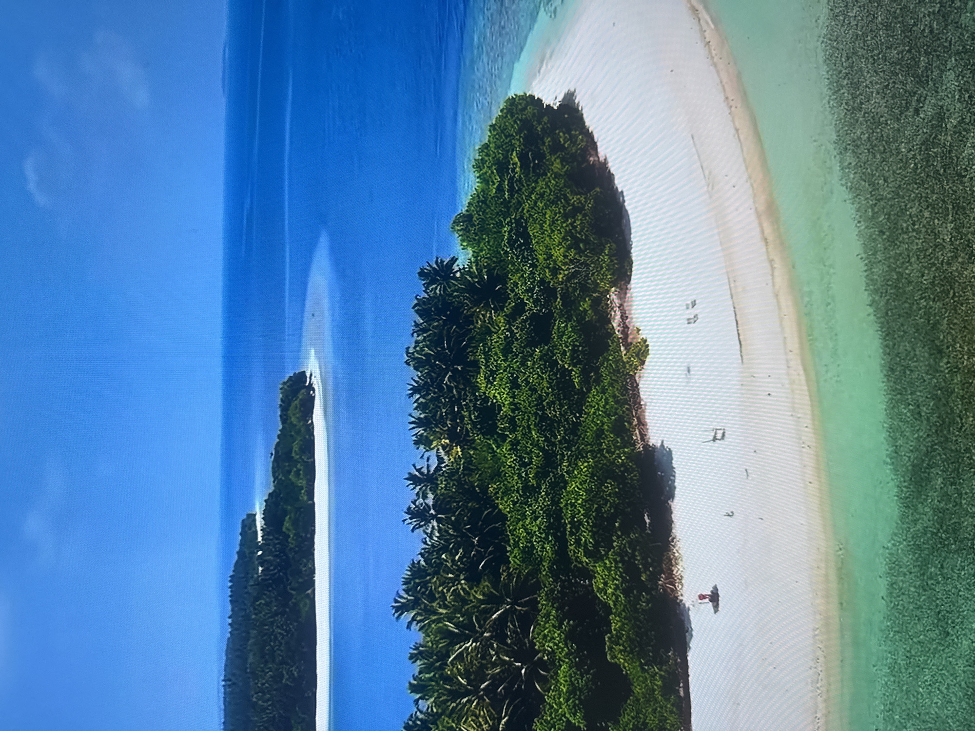 The image is an aerial view of a tropical island landscape. The foreground features a white sandy beach that curves into the turquoise water. The water transitions to a deeper blue as it recedes into the distance. Lush green vegetation, primarily palm trees, covers the island. In the mid-ground, another island or sandbar with similar vegetation is visible, separated by the blue ocean. The sky is clear and bright blue. There are a few small, indistinct objects on the beach, one appearing to be a person in red clothing and two chairs.
This image does not depict the city of Almaty, Kazakhstan. It is a natural tropical setting.