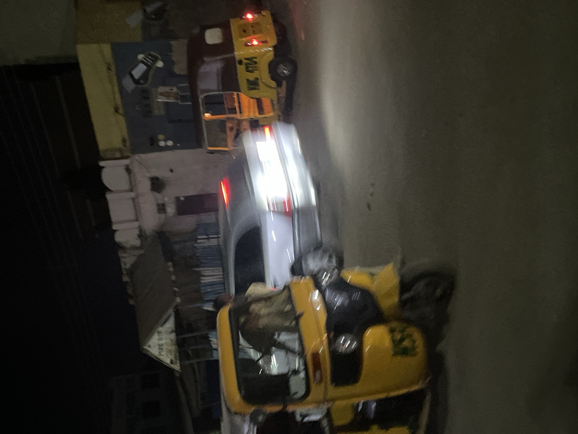 Here's a detailed description of the image:
**Scene:** A street in an unidentified city, likely at night or in low-light conditions.
**Objects:**
* **Vehicles:** Several vehicles are visible. One appears to be a white car or small truck with headlights on. Two yellow tuk-tuks (auto-rickshaws) are also present.
* **Buildings:** Buildings of various heights line the street.
* **Other objects:** A pile of books.
**People:**
* **Person on a tuk-tuk:** A person, possibly wearing dark clothing, is seated on one of the tuk-tuks.
**Environment:**
* **Light:** The overall lighting is poor, making it difficult to discern fine details. There are street lights and vehicle headlights.
* **Setting:** The scene suggests an urban setting with a mix of commercial and residential buildings.