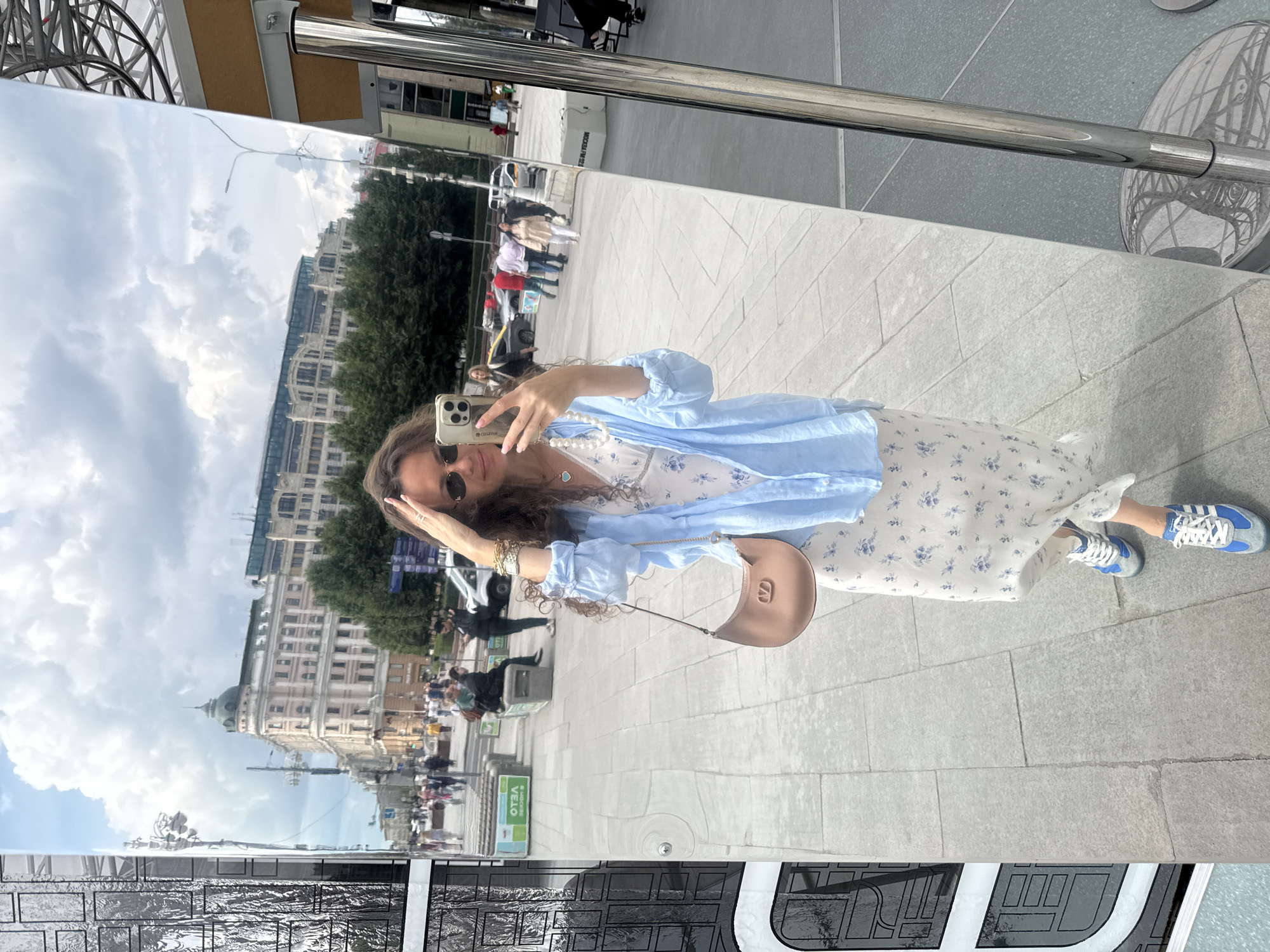 A young woman with long, curly brown hair takes a selfie in a large, full-length mirror. She is wearing sunglasses, a light blue unbuttoned shirt over a white floral-print dress, a light brown handbag with a gold 'V' logo, a pearl necklace, and blue and white Adidas sneakers. In the background, reflected in the mirror, is a city street scene with buildings, trees, people walking, and cars. The architectural style of the buildings suggests a location in Moscow, Russia. The woman is standing on a paved walkway with square stone tiles. The mirror is mounted on a chrome pole.