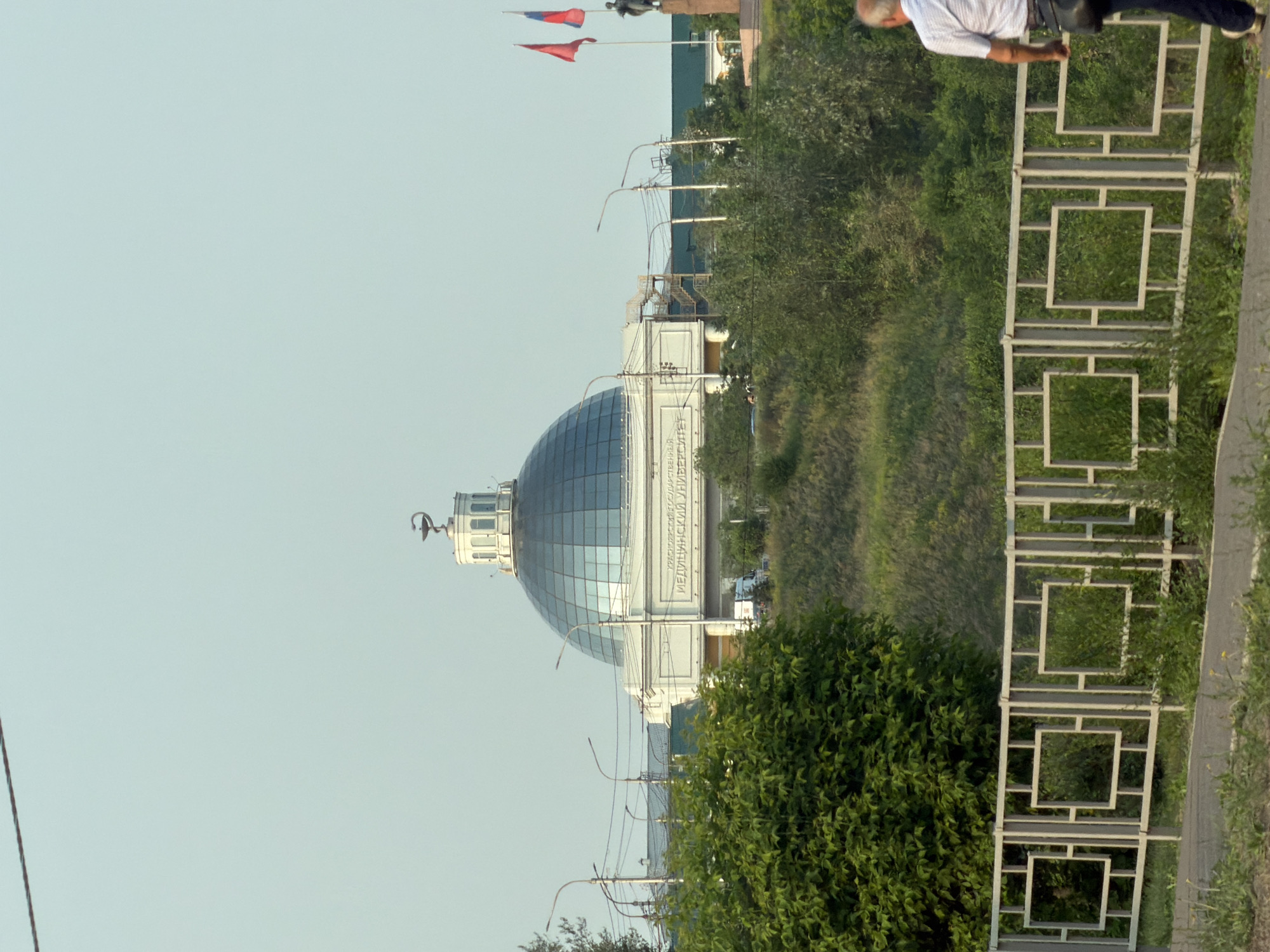 The image depicts the Krasnoyarsk State Medical University in Krasnoyarsk, Russia, featuring its distinctive dome topped with a caduceus symbol. The university building, with its classical facade and large glass dome, is situated on a grassy embankment. Two flags, one red and one red and blue, fly on flagpoles near a statue to the right of the building. A white metal fence with square geometric patterns runs along the foreground, separating a paved path from the embankment. Green foliage, including a large tree on the left, covers parts of the embankment. In the background, overhead wires and streetlights are visible. A person, a male with gray hair wearing a light-colored shirt and dark pants, walks away from the camera along the path. The sky is clear and light blue.