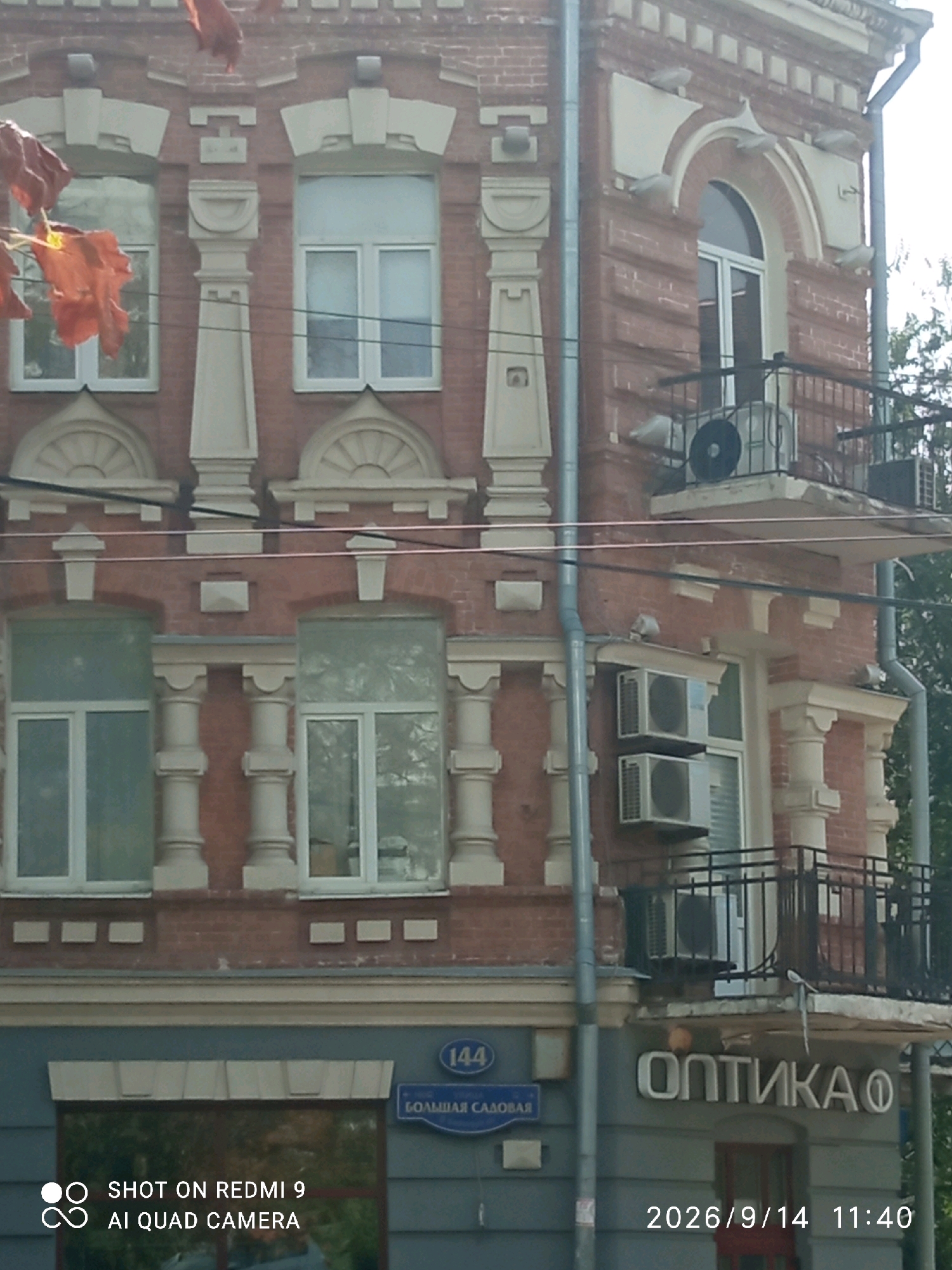This is an exterior, eye-level, medium shot of a multi-story brick building in Rostov-on-Don, Russia. The building features ornate architectural details, including pilasters, decorative molding, arched windows, and balconies with railings. Several windows are visible, some with air conditioning units mounted outside. On the lower level, a sign reads "ОПТИКА" (Optics) in large, white, sans-serif letters with a small circle to the right. Above the sign, there is a street sign with "144" and "БОЛЬШАЯ САДОВАЯ" (Bolshaya Sadovaya) written on it. The timestamp "2026/9/14 11:40" is visible in the bottom right corner, along with the text "SHOT ON REDMI 9 AI QUAD CAMERA" in the bottom left. The scene appears to be during daylight hours, with trees visible in the background.