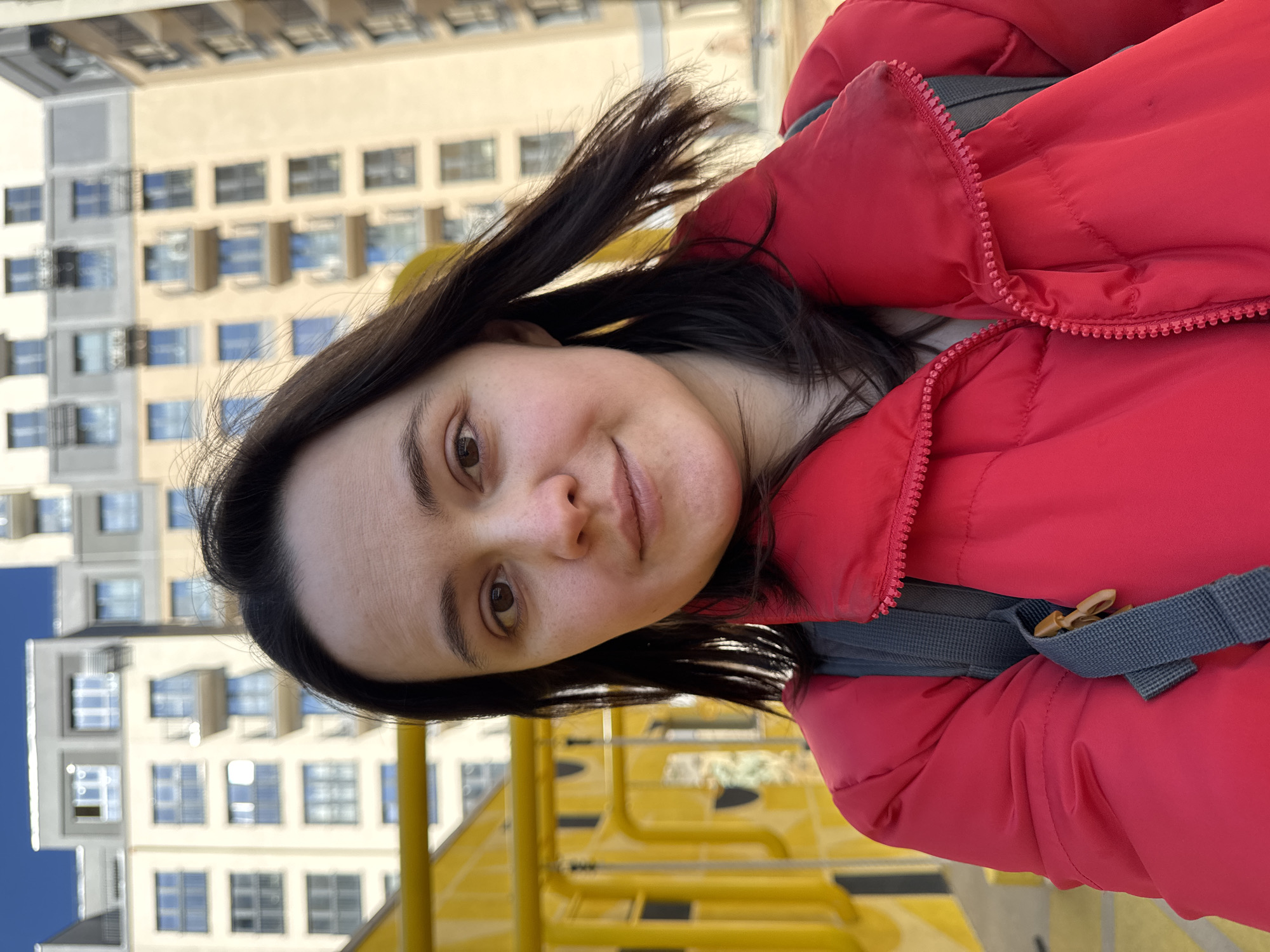 A woman is the subject of a medium shot, angled from below. She has dark hair and brown eyes. She wears a red puffy jacket with gray straps over her shoulders. Behind her is a bright yellow metal structure, likely a playground element. In the background stands a tall building with many windows, consistent with apartment architecture in Izhevsk, Russia. The sky is visible in the upper left corner, appearing blue.