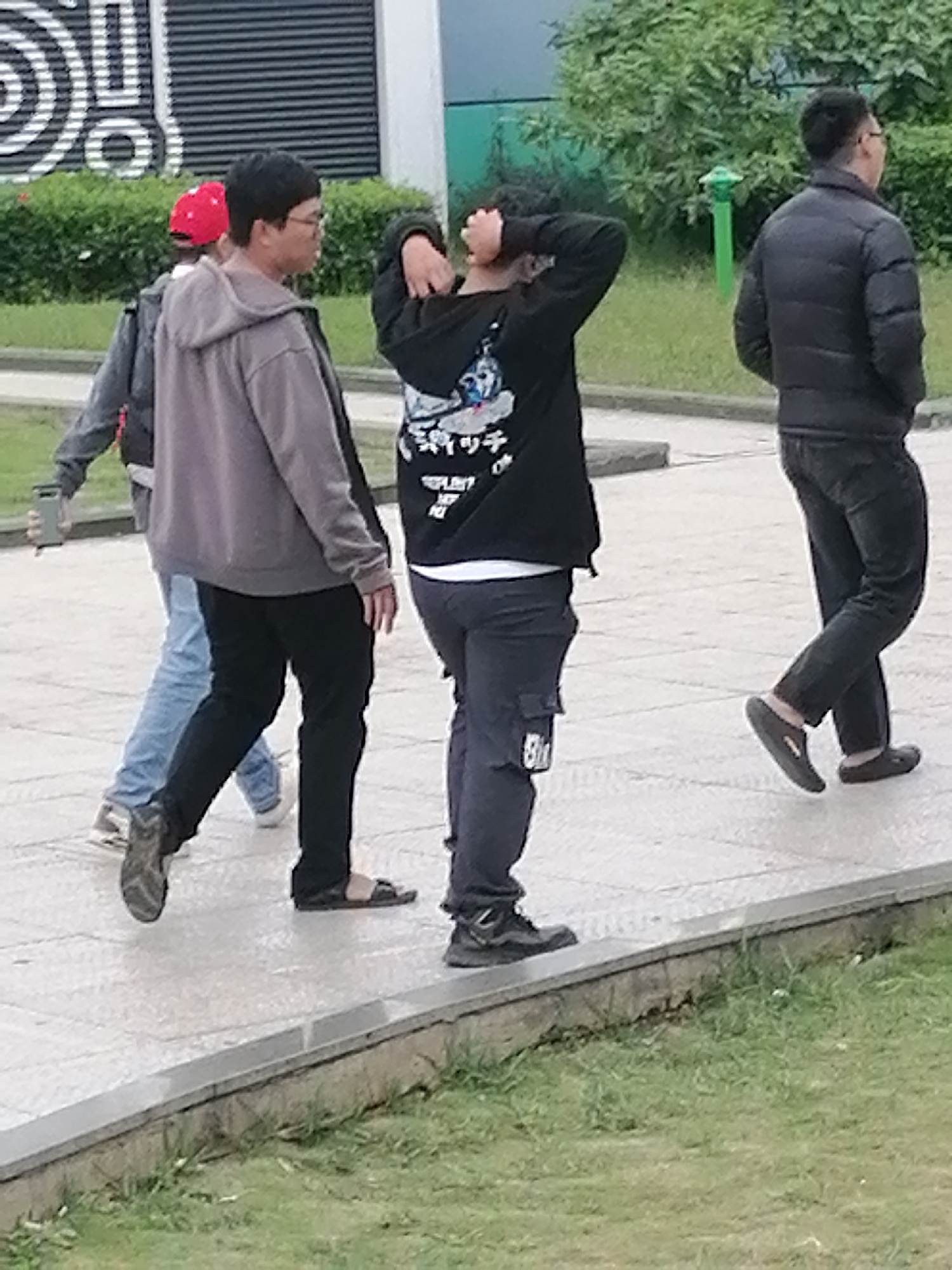 The image shows four people walking on a paved pathway. The person in the foreground on the left is wearing a grey hooded sweatshirt, black pants, and black sandals. To their right, another person is wearing a black hooded sweatshirt with graphics on it, dark pants, and black shoes. To the left of the first person, a person in a red cap and grey jacket is walking. Behind them, a person in a black puffer jacket and dark pants is walking. The background shows green bushes and a building with horizontal dark grey blinds. The scene appears to be outdoors in an urban or park-like setting. There are no clear indicators of the specific location within Da Lat, Vietnam.