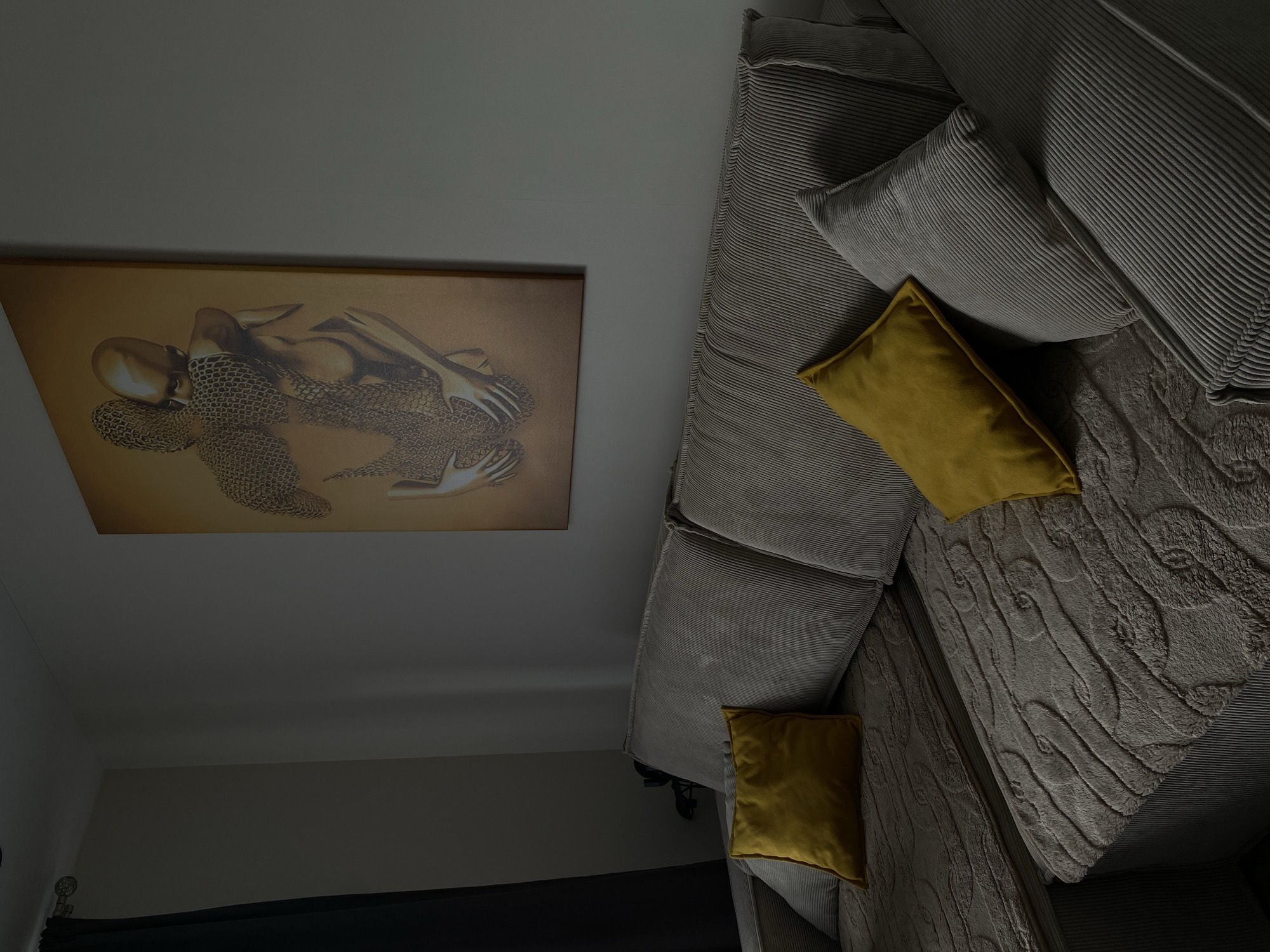 An indoor scene showcases a comfortable living space, centered around a large, light grey corduroy sectional sofa. The sofa is furnished with several matching grey corduroy cushions and two vibrant mustard yellow throw pillows, providing a pop of color. A textured, light grey throw blanket, possibly knitted or embossed with a cable-knit pattern, drapes over a portion of the sofa. On the wall above the sofa, a large framed artwork is prominently displayed. This artwork features a stylized, golden figure or figures, depicted with a distinctive chainmail-like texture. The walls and ceiling are a uniform light grey. In the lower left corner, a dark curtain rod with a decorative finial and dark curtains are partially visible, suggesting a window or doorway. The lighting in the room is dim and appears to be artificial, indicating an indoor setting, possibly during the evening or night. No people or specific activities are depicted in the static image. No text is visible.