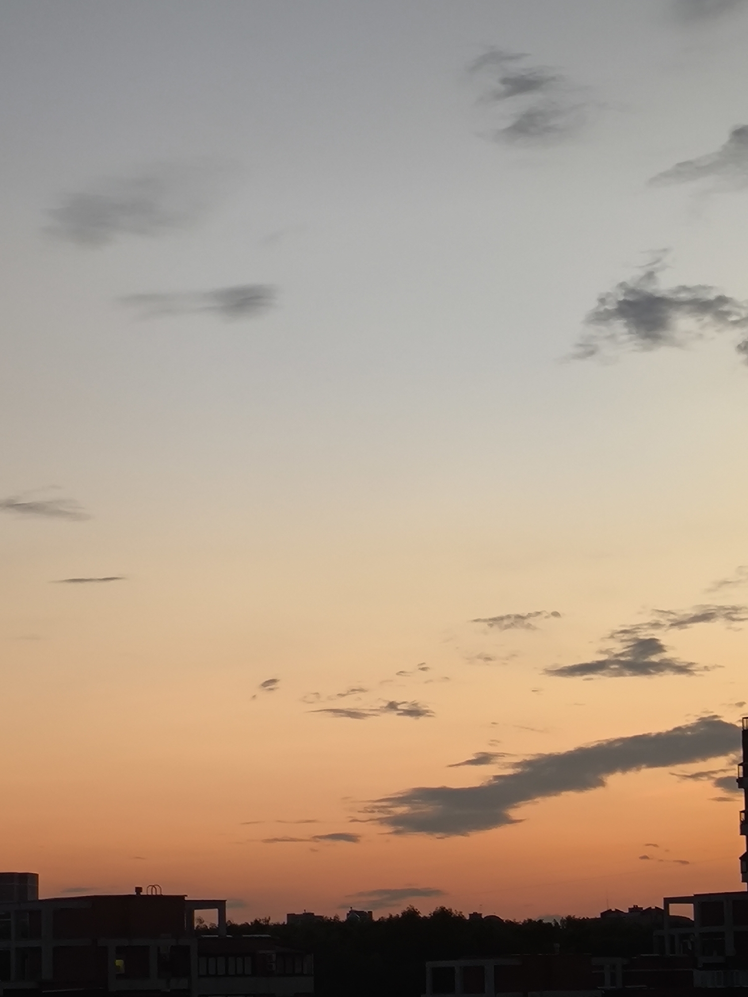 The image shows a sky with clouds at sunset. The sky transitions from a light blue at the top to a warm orange near the horizon. There are scattered clouds throughout the sky, some appearing dark and wispy, others lighter and more diffused. In the lower portion of the image, the silhouettes of buildings and trees are visible against the colorful sky. The architecture suggests an urban or suburban setting.