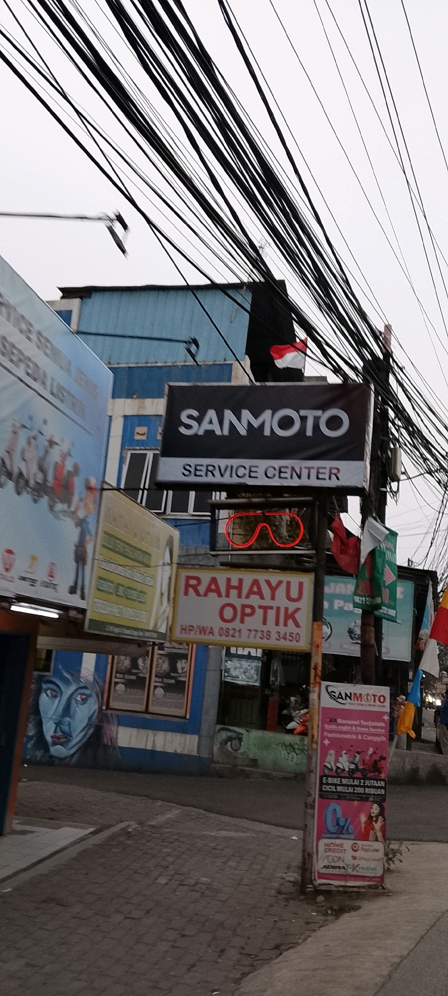 The image captures a street view in Depok, Indonesia, characterized by a dense network of overhead electrical wires against a pale sky. In the foreground, a paved sidewalk and road are visible. Several signs are mounted on poles and buildings. A prominent black sign with white lettering reads "SANMOTO SERVICE CENTER." Below this, a red neon sign shaped like eyeglasses glows. Another sign, oriented vertically, displays "RAHAYU OPTIK" in large red letters with a phone number in smaller text below. To the left, a large blue and white billboard features images of motorcycles and people. Further down the street, other signs and building facades are partially visible, along with graffiti on a wall depicting a face. The overall scene suggests a commercial area with multiple businesses.