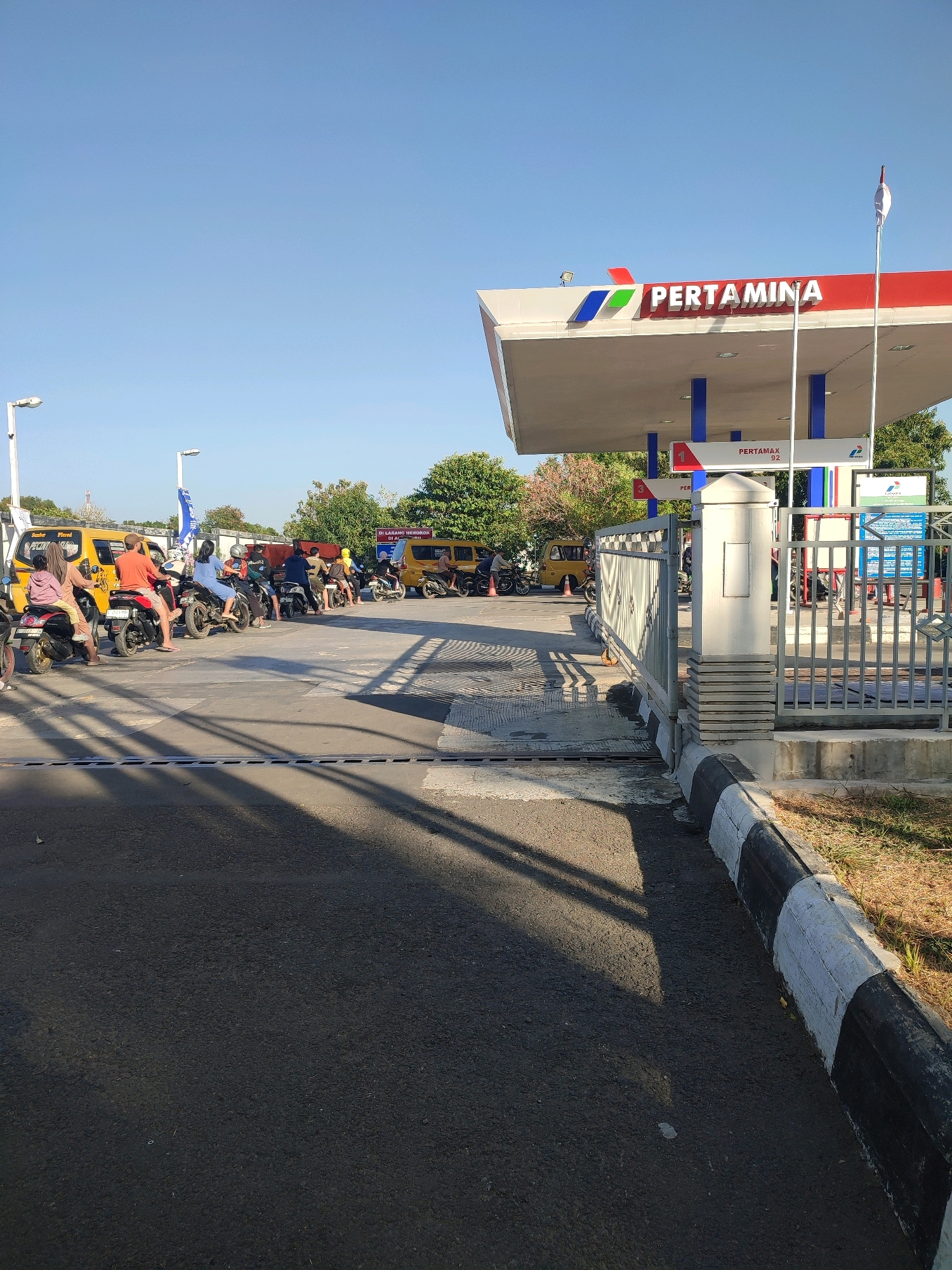 The image depicts a Pertamina gas station in Watubelah, Indonesia, on a clear, sunny day. Several motorcycles and a yellow van are queued at the pumps. A person in an orange shirt is riding a motorcycle in the front of the line, while another person in a pink hoodie is seated behind them. The Pertamina logo is prominently displayed on the canopy of the gas station, with "PERTAMAX 92" visible on a sign. A white flag with a red stripe flies from a pole near the canopy. A metal fence encloses part of the gas station property. The ground is asphalt, with a painted curb along the edge. Trees and foliage are visible in the background, and shadows are cast across the pavement.