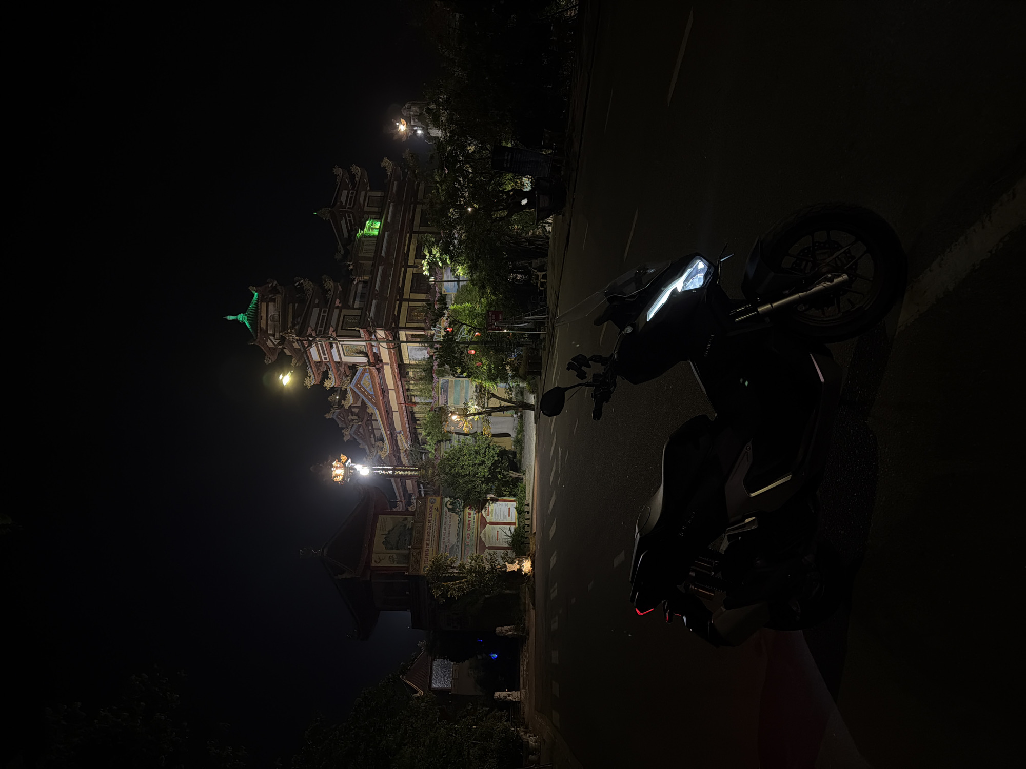 Captured at night in the city of Đà Nẵng, Vietnam, the image features a motorcycle parked on the side of a road. In the background, a brightly illuminated and intricate Buddhist temple or architectural structure is visible. Street lamps are scattered around, contributing to the overall lighting of the scene. A sign with the text "CHUA QUAN THE AM NGU QUẠC ĐAI SEN NGOC LEHOI LE HOI QUAN THE DONON" is partially visible.