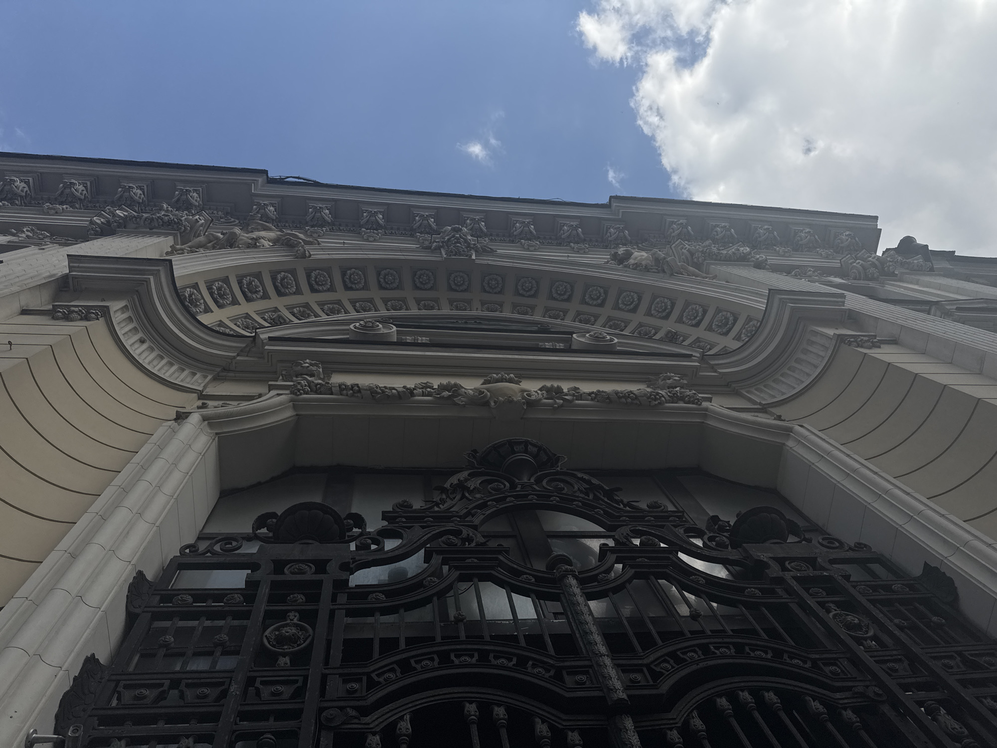 Low-angle view of an ornate building facade in Moscow, Russia. The building is beige with elaborate gray and white architectural details. The roofline is decorated with carvings. The facade features a large, decorative black iron window grille. The sky is blue with scattered white clouds. No people are visible.