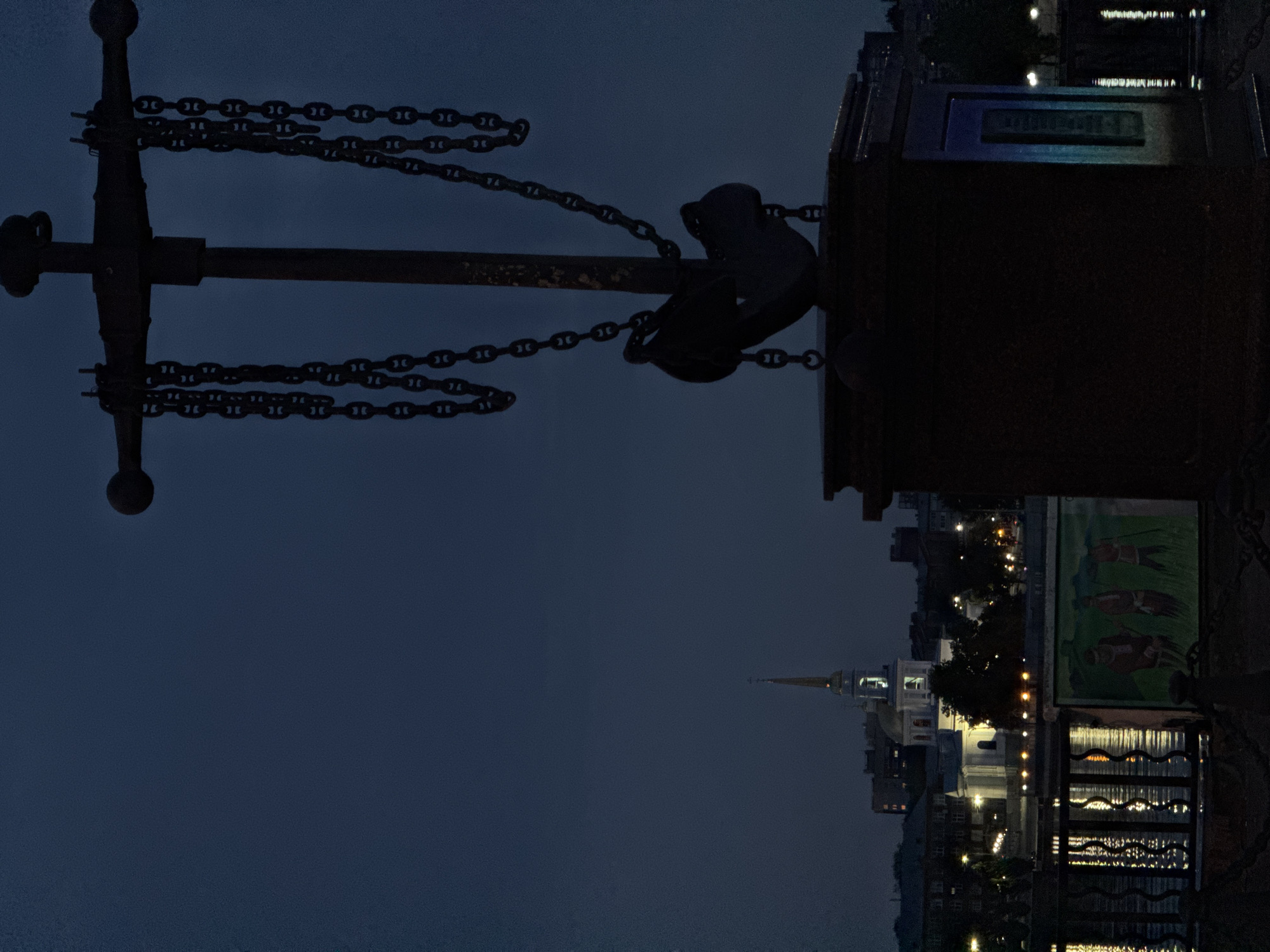 A large, dark anchor, attached to heavy chains, dominates the foreground of the image, silhouetted against a twilight sky. The anchor sits atop a dark, rectangular pedestal. In the background, across a body of water reflecting lights, the city of Votkinsk, Russia, is visible. A white church with a tall, golden spire stands out against the darker buildings. The water is rippled and reflects the lights from the city and possibly from the streetlights along the embankment. A mural depicting three figures in traditional clothing is visible on a barrier in the middle ground. The overall atmosphere is serene and somewhat somber, suggesting early evening or dawn.