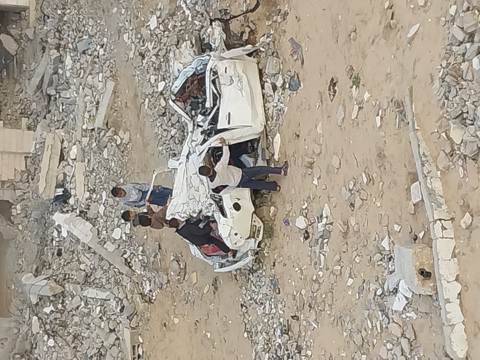 Five children are depicted interacting with a severely damaged white passenger vehicle amidst extensive rubble in Khan Yunis, Palestinian Territory. The vehicle, appearing to be a sedan, is crushed, distorted, and partially inverted, with its roof collapsed and interior components exposed. The surrounding ground consists of light brown dirt heavily interspersed with grey concrete blocks, broken masonry, and rebar, indicating widespread structural destruction. In the background, large piles of similar debris and remnants of demolished buildings extend across the landscape. One child in a light grey hoodie stands to the left of the car, observing it. Another child in a brown jacket stands near the vehicle's crumpled roof. A third child in a dark jacket is positioned on the left side of the car, seemingly climbing on it. A fourth child, wearing a light-colored long-sleeved shirt and dark pants, stands by the open right-side door, holding onto its frame. A fifth child is partially obscured behind the brown-jacketed child. In the foreground, a broken concrete curb or foundation element is visible. The scene is illuminated by natural daylight.