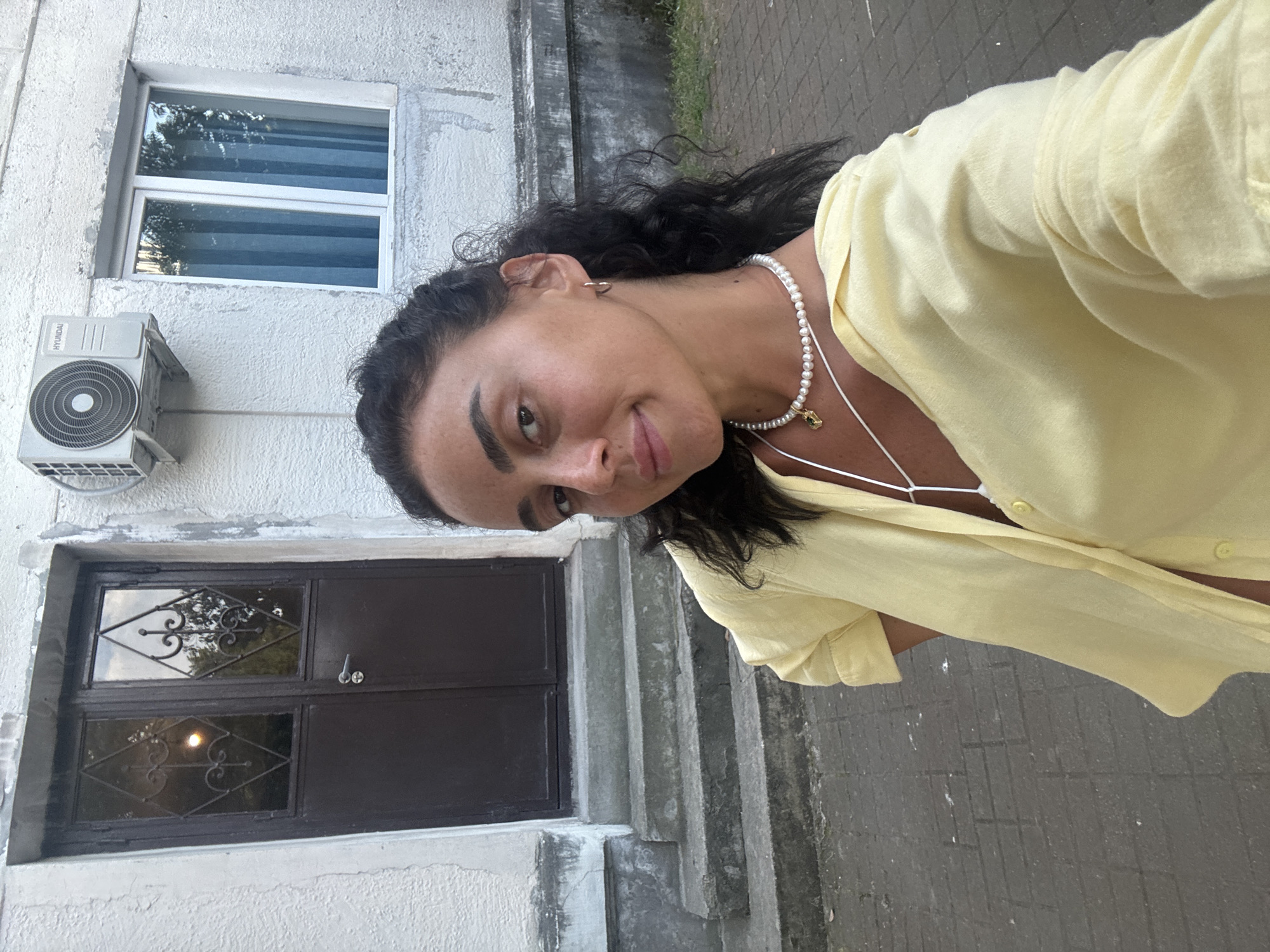 In Batumi, Georgia, a woman with dark curly hair and fair skin, wearing a yellow button-up shirt over a white top and a pearl necklace with a pendant, is taking a selfie outdoors. She is positioned in front of a building with a brown double door entrance, concrete stairs, a window with blue curtains, and an air conditioning unit mounted on the wall. The ground is paved with brick-like stones.