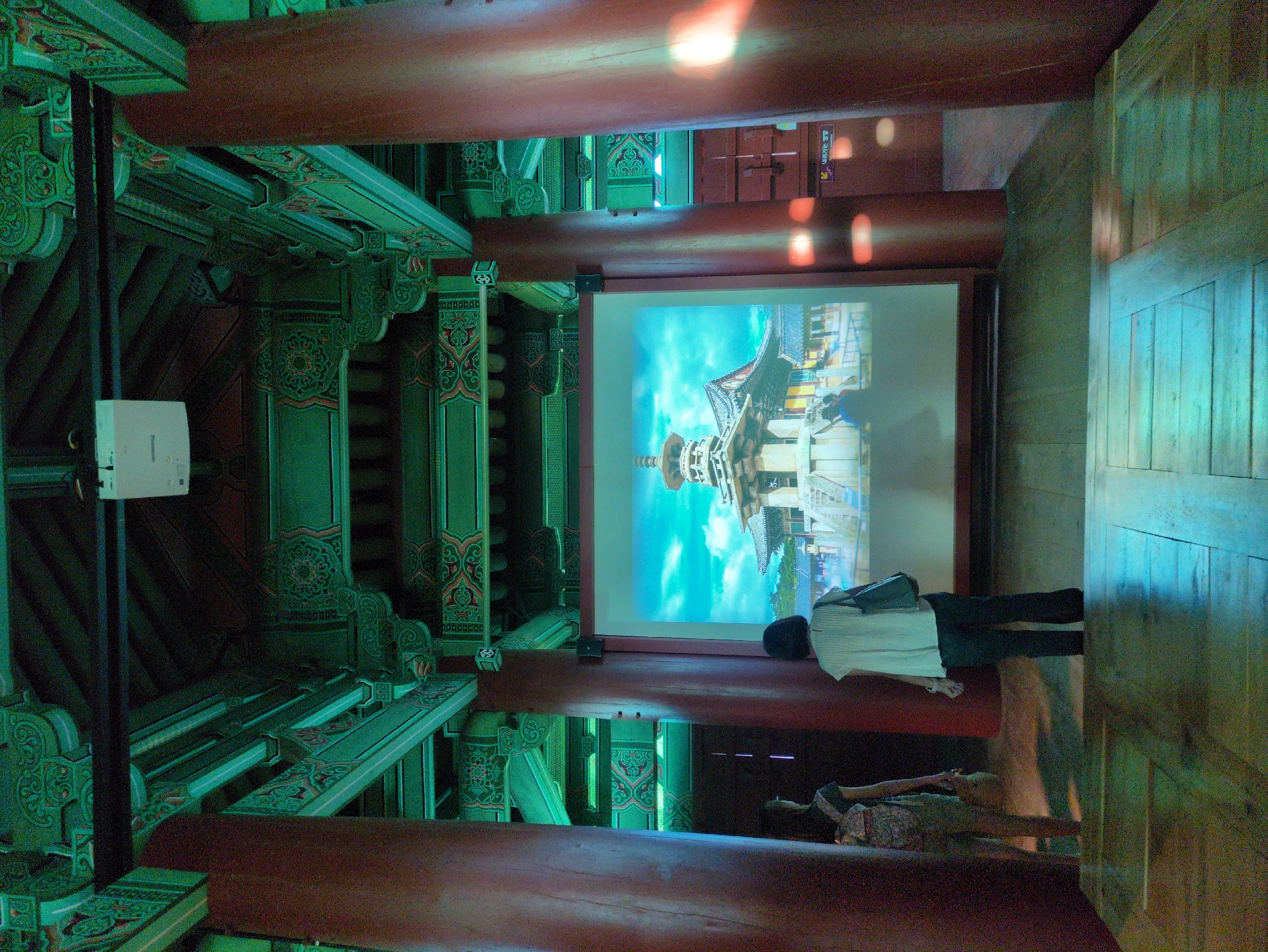 The image depicts an interior view of a structure in Gyeongju-si, South Korea. The architecture features ornate green and red painted wood with intricate patterns. A projector mounted on the left side of the frame projects an image of what appears to be a pagoda or multi-tiered tower onto a screen in the center. Two individuals are present. One, dressed in a light-colored shirt and dark pants, stands in front of the screen. The other individual, wearing a patterned dress, is seated on a red wooden beam. The floor is also made of wood, with a bluish hue suggesting artificial lighting.