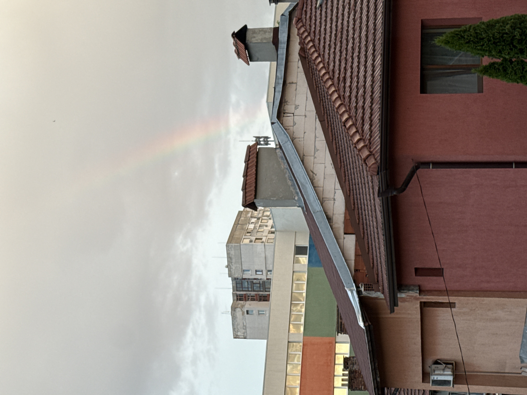 The image captures an overcast sky over a city landscape, with a faint rainbow visible in the upper portion of the frame. In the foreground, the tiled roofs of residential buildings dominate the view. One building features a terracotta-colored facade with a visible window and a small evergreen tree in front of it. Another building to the left is a lighter shade, with an air conditioning unit mounted on its exterior wall. Behind these, a larger, more modern building with a colorful facade, featuring bands of orange, green, and blue, is visible. Further in the background, a tall, grey concrete apartment building with numerous windows stands against the sky. The scene appears to be set in an urban environment, possibly a residential neighborhood within Sofia, Bulgaria, given the architectural styles of the buildings and the presence of apartment blocks commonly found in Eastern European cities. There are no people or animals visible in the image. The overall impression is one of a calm, post-rain city scene.