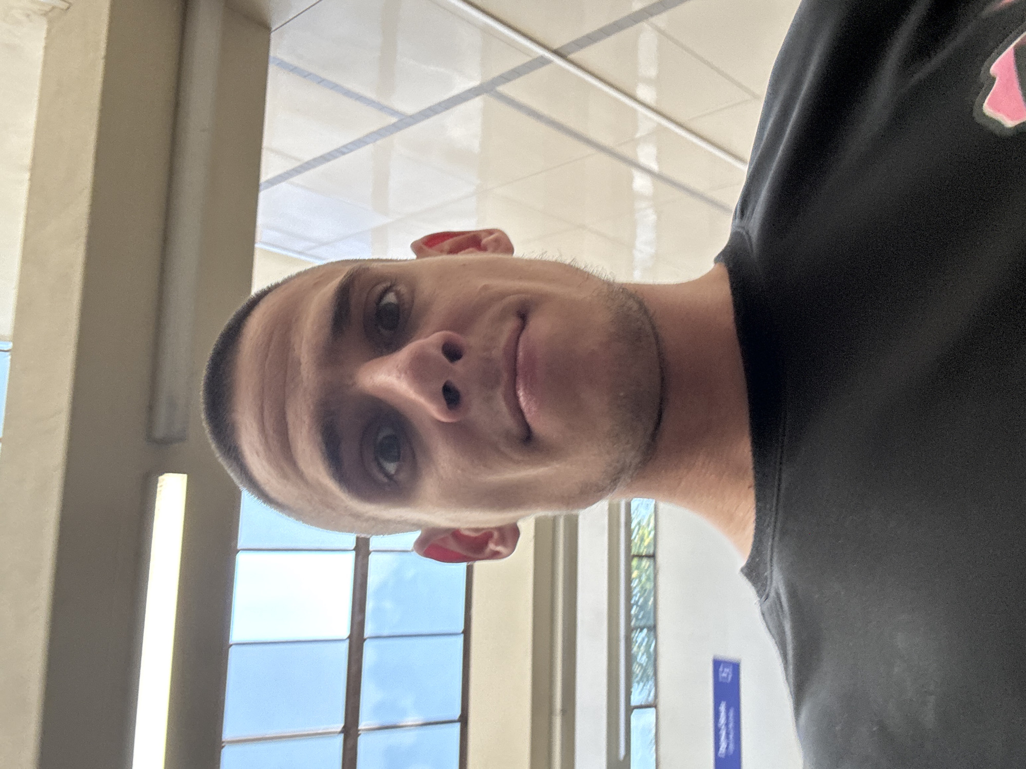 The image is a close-up selfie of a man with a buzz cut, fair skin, and brown eyes. He is wearing a black t-shirt with a pink graphic design on the lower right side, partially visible. The man is looking directly at the camera with a neutral expression. The background suggests an indoor location with large windows or skylights showing a blue sky with clouds. There are fluorescent lights visible above the windows. Further in the background, there are architectural elements that appear to be part of a modern building, possibly a public transport station or a large hall, with geometric patterns and a cream-colored ceiling. A blue sign with white text is visible on the left side of the image, near the man's shoulder. Based on contextual information provided in the prompt, this scene is located in Sofia, Bulgaria, likely within a public transportation hub such as a metro station or bus terminal.