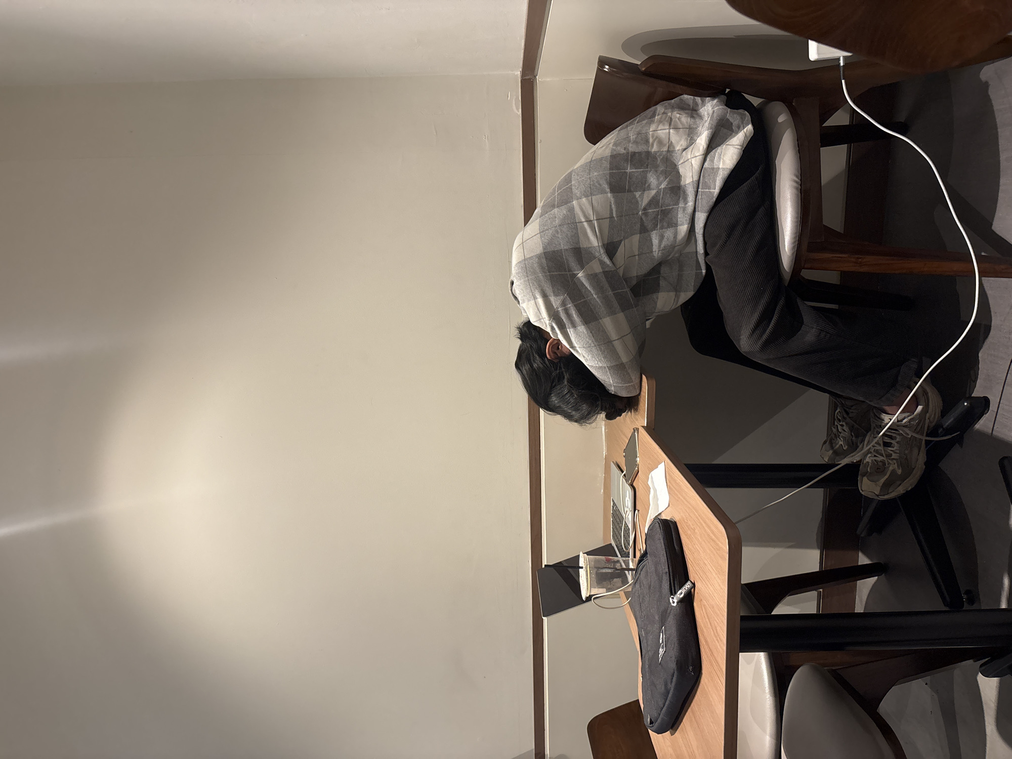 A person is sleeping with their head on a wooden table in a dimly lit room, possibly a cafe or study area, with a laptop, a cup with a straw, a phone, and a black laptop bag on the table. The person is wearing a grey and white checkered shirt and dark pants, and their legs are crossed with sneakers on. White power cords are visible running across the floor and on the table. The background consists of a plain wall with a horizontal wooden trim and a spotlight illuminating a section of the wall. The overall impression is one of exhaustion or prolonged study. The location is not specified in the image, and therefore cannot be determined as West Jakarta, Indonesia.