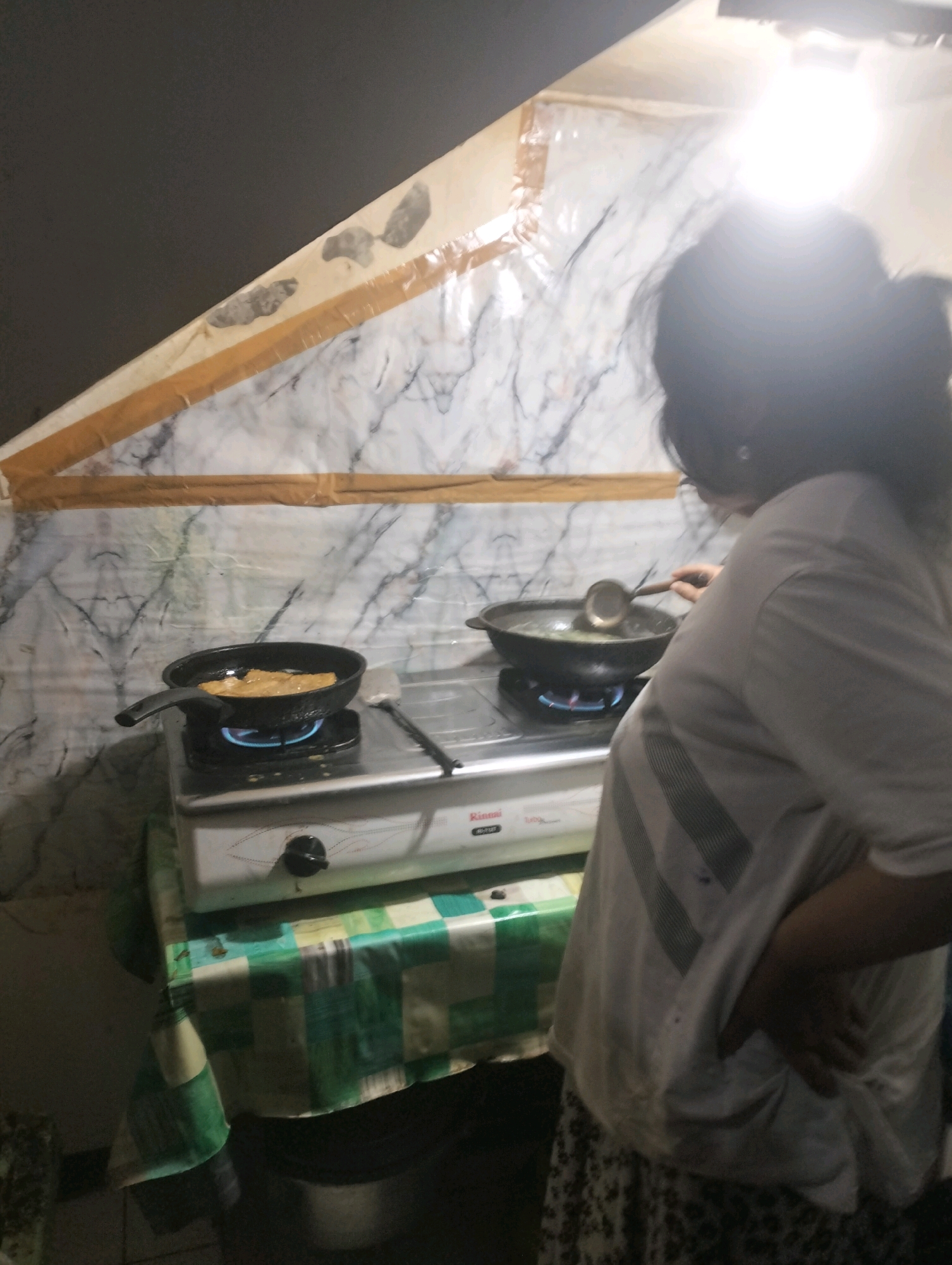 In Klecorejo, Indonesia, a woman is cooking on a portable gas stove. The stove is positioned on a table covered with a green and white checkered tablecloth. The background features a wall covered in a marble-patterned wallpaper, taped to a slanted ceiling. A bright light is illuminating the scene from above, casting shadows and creating a high-contrast environment. The woman, seen from the side and slightly from behind, is wearing a white t-shirt and a patterned skirt. She is actively engaged in cooking, using a ladle to stir something in a pan on the stove. Another pan with food is visible on a different burner of the stove. The overall impression is of a simple, functional cooking setup in a home environment.