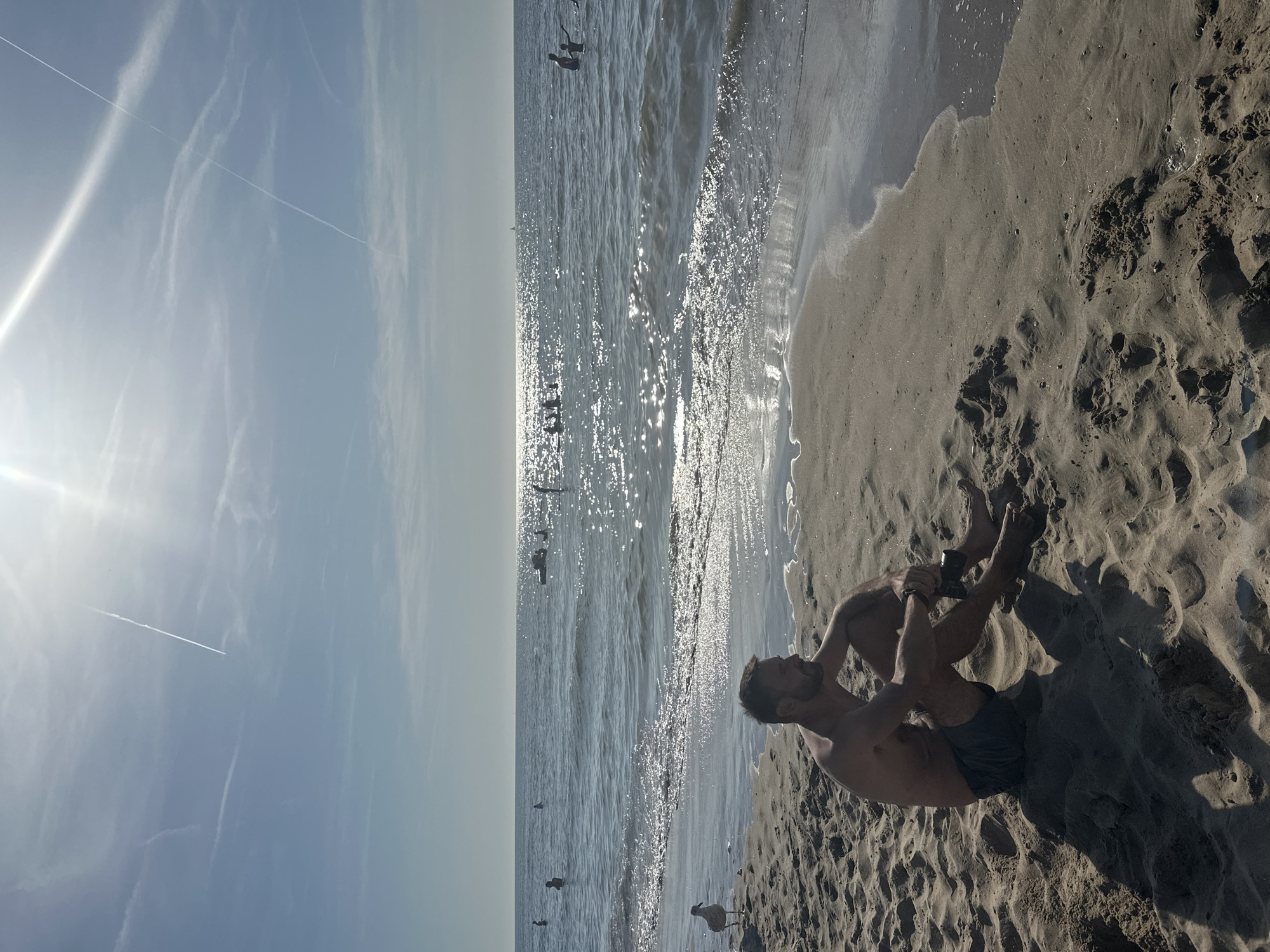 A man is sitting on the sand at the beach in Zandvoort, Netherlands. He is facing to his right, with his legs crossed and his arms resting on his knees. He is wearing dark swim trunks. The sun is shining brightly, and there are some contrails in the sky. The sea is calm, with a few people swimming in the distance. A seagull is standing on the sand to the left of the man. The sand is wet near the water's edge and dry further up the beach.