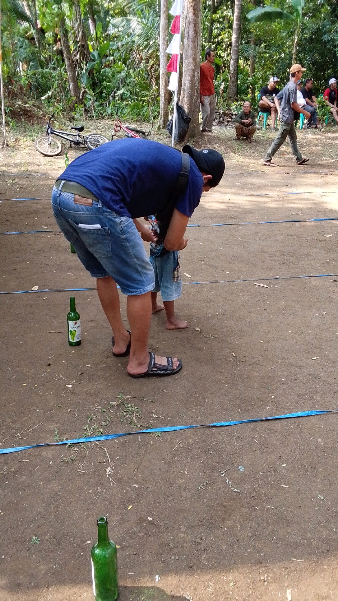In a dusty, outdoor area, likely in Parakanmanggu, Indonesia, a man wearing a blue t-shirt and denim shorts bends over, interacting with a child. The man has a black baseball cap on and is wearing sandals. The child, wearing a plaid patterned shirt and shorts, stands barefoot. To the man's left, a green bottle with a label featuring grapes stands upright on the ground. Further in the background, several men are seated on plastic chairs, and others are standing. One man in a grey t-shirt and green pants walks towards the right of the frame. Trees and foliage form the backdrop, with a red and white flag visible on a pole. Two bicycles are lying on the ground in the left background. A blue ribbon runs horizontally across the foreground, marking a boundary. Another green bottle lies on its side in the lower-left corner of the image.