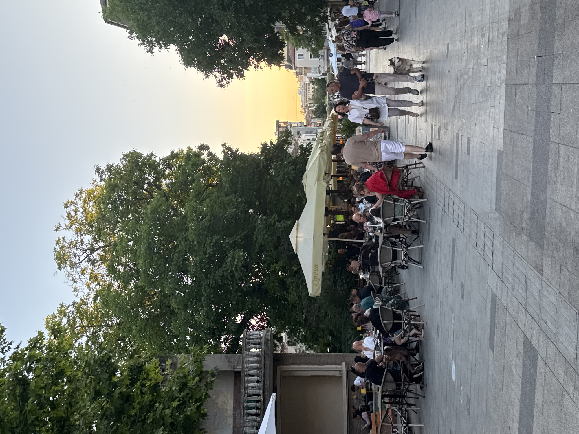 The image depicts a street scene in Burgas, Bulgaria, likely during the evening. In the foreground, a paved walkway is lined with outdoor seating for restaurants and cafes. People are seated at tables, some dining, others conversing. A couple walks hand-in-hand down the street, accompanied by a husky dog. Further down the street, more pedestrians are visible, along with buildings and trees. The sky shows a gradient of colors, suggesting sunset or sunrise. The atmosphere appears relaxed and social.