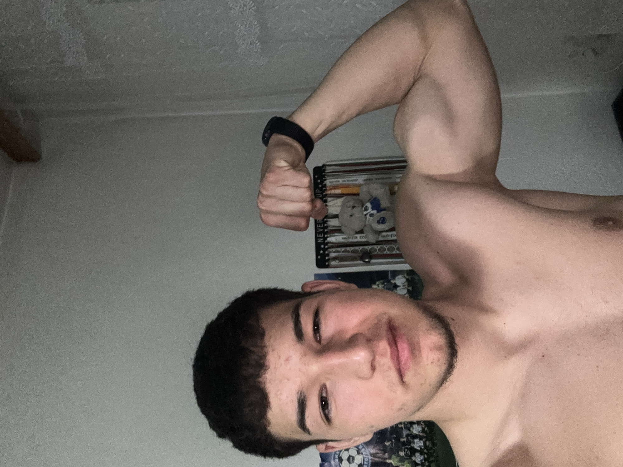 A shirtless young man poses with his right arm flexed in an indoor setting. He wears a black fitness tracker on his wrist. The background shows a wall with a patterned wallpaper, and a shelf containing books and stuffed animals. No evidence suggests this image was taken in Zlatograd, Bulgaria.