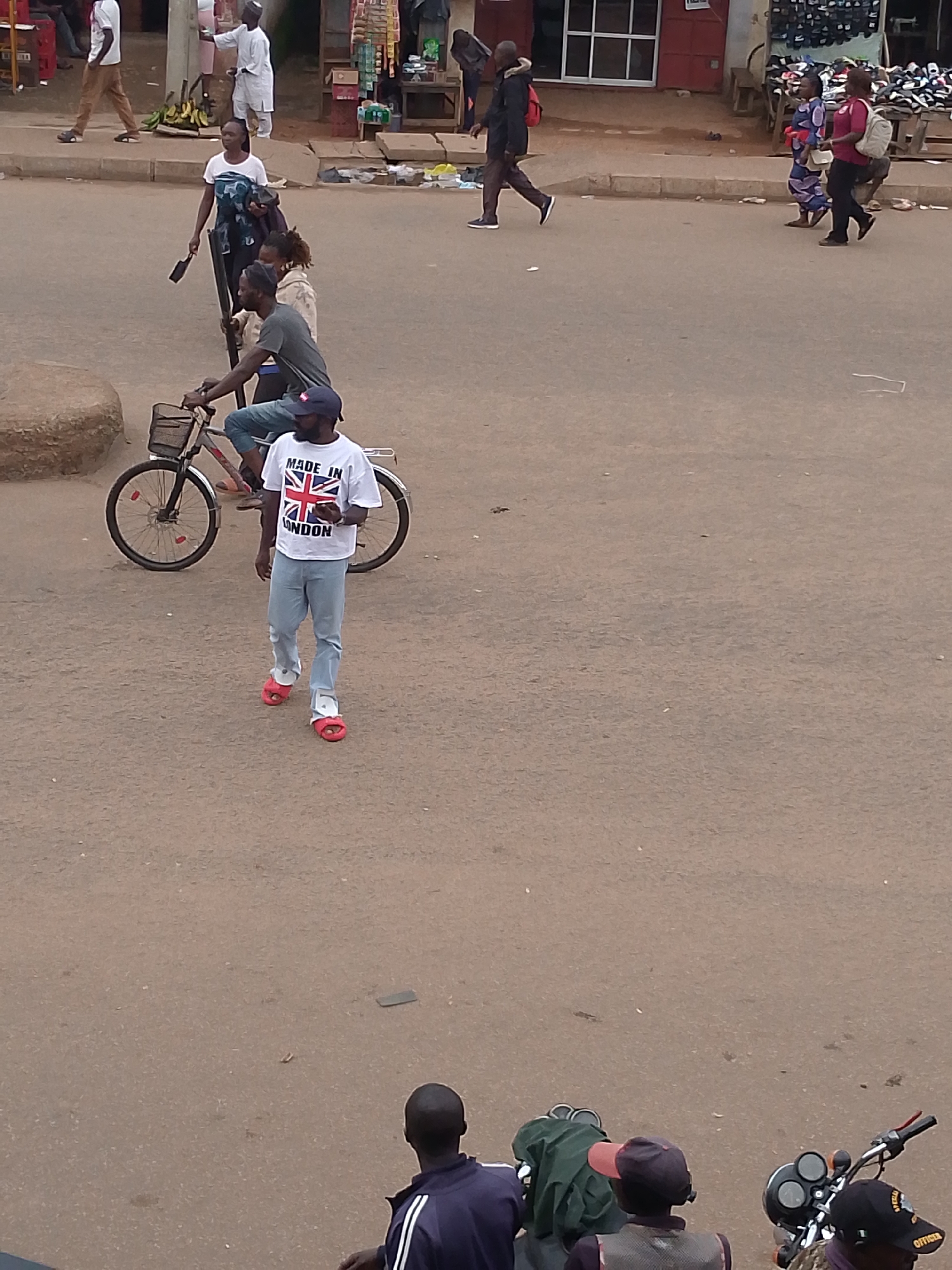 In Bukuru, Nigeria, a street scene unfolds with multiple individuals. In the foreground, a man wearing a white t-shirt with the Union Jack and the words "MADE IN LONDON" walks on roller-skates. He is accompanied by a man on a bicycle. Further in the background, several other people are walking, including a woman in a white t-shirt and a woman in a blue and white patterned dress. The environment appears to be a public street with shops visible in the distance, suggesting a marketplace or commercial area. The ground is paved, and there are some debris scattered on the road.