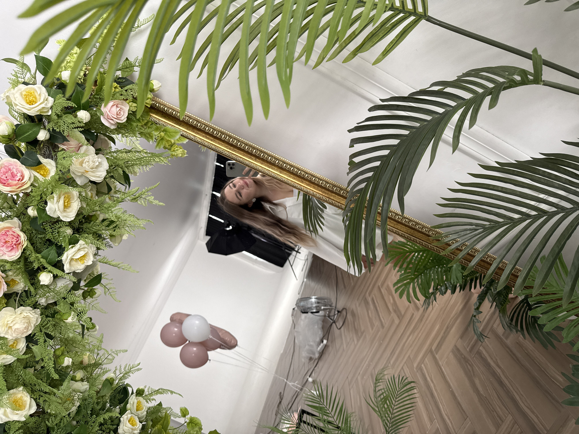 A mirror, framed with gold and surrounded by artificial flowers and palm leaves, reflects a woman taking a selfie. The reflection shows a light-skinned woman with long brown hair, wearing a white top, holding a phone to capture her image. Behind her in the reflection is a room with white walls, a herringbone pattern floor, and balloons. A studio light is also visible. The location is likely indoors, and could be a studio or a room decorated for an event. The information available does not confirm it being in the city of Chaikovsky, Russia.