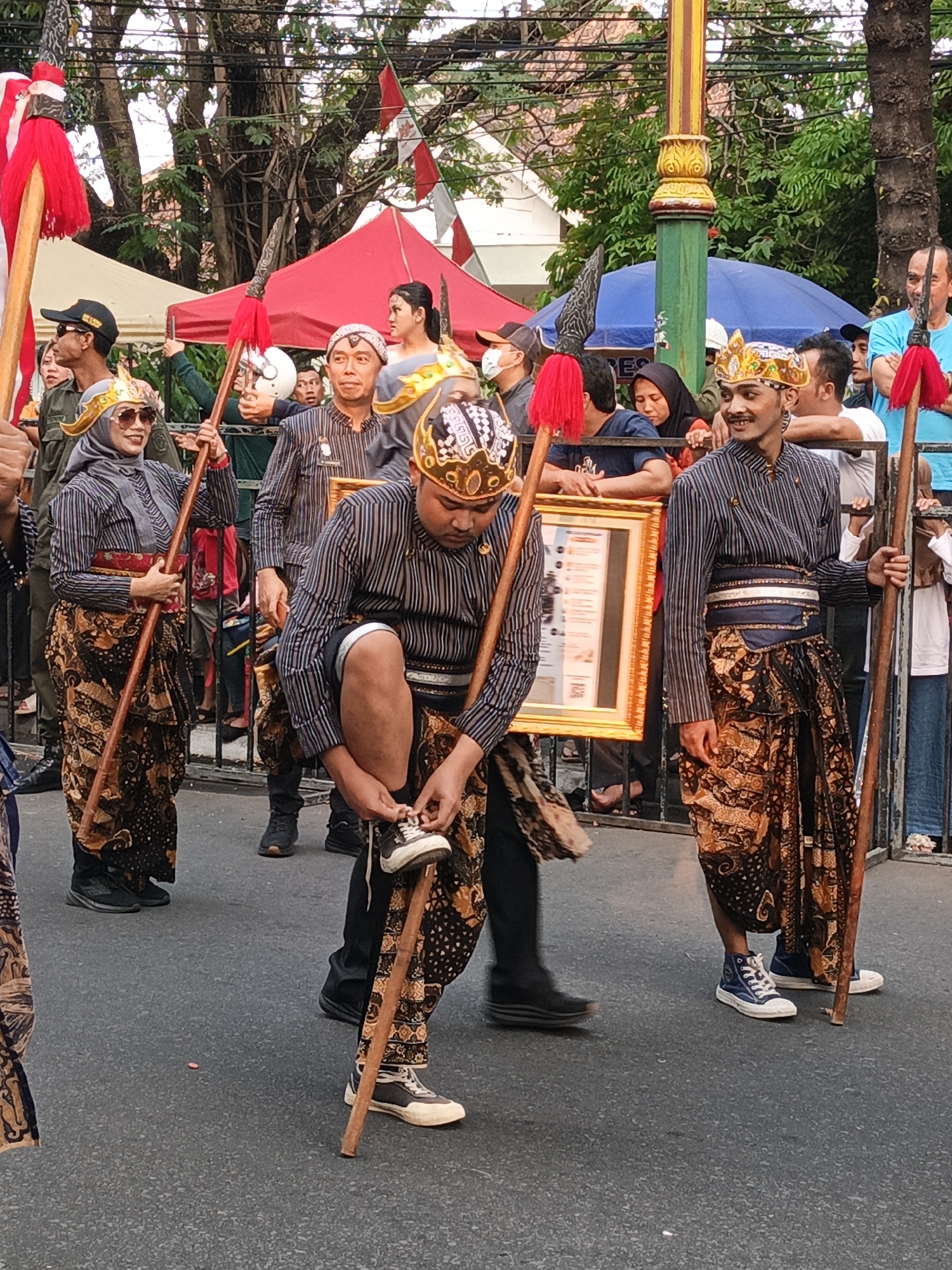 In Gayamprit, Indonesia, a procession is underway. A man, dressed in traditional Javanese attire including a striped long-sleeved shirt, a batik sarong, and a golden headdress, is seen kneeling to tie his shoelaces. He holds a spear with a red tassel in his left hand and another spear with a red tassel stands next to him on his right. To his left, a woman, also in traditional Javanese clothing with a grey headscarf and sunglasses, holds a spear with a red tassel. She wears a striped long-sleeved top and a batik sarong. Behind them, a framed document with Asian writing is held by someone out of frame. Other individuals in similar traditional attire and modern clothing are visible in the background, some holding spears. Tents and trees line the street, with onlookers standing behind a barrier. The ground is asphalt.