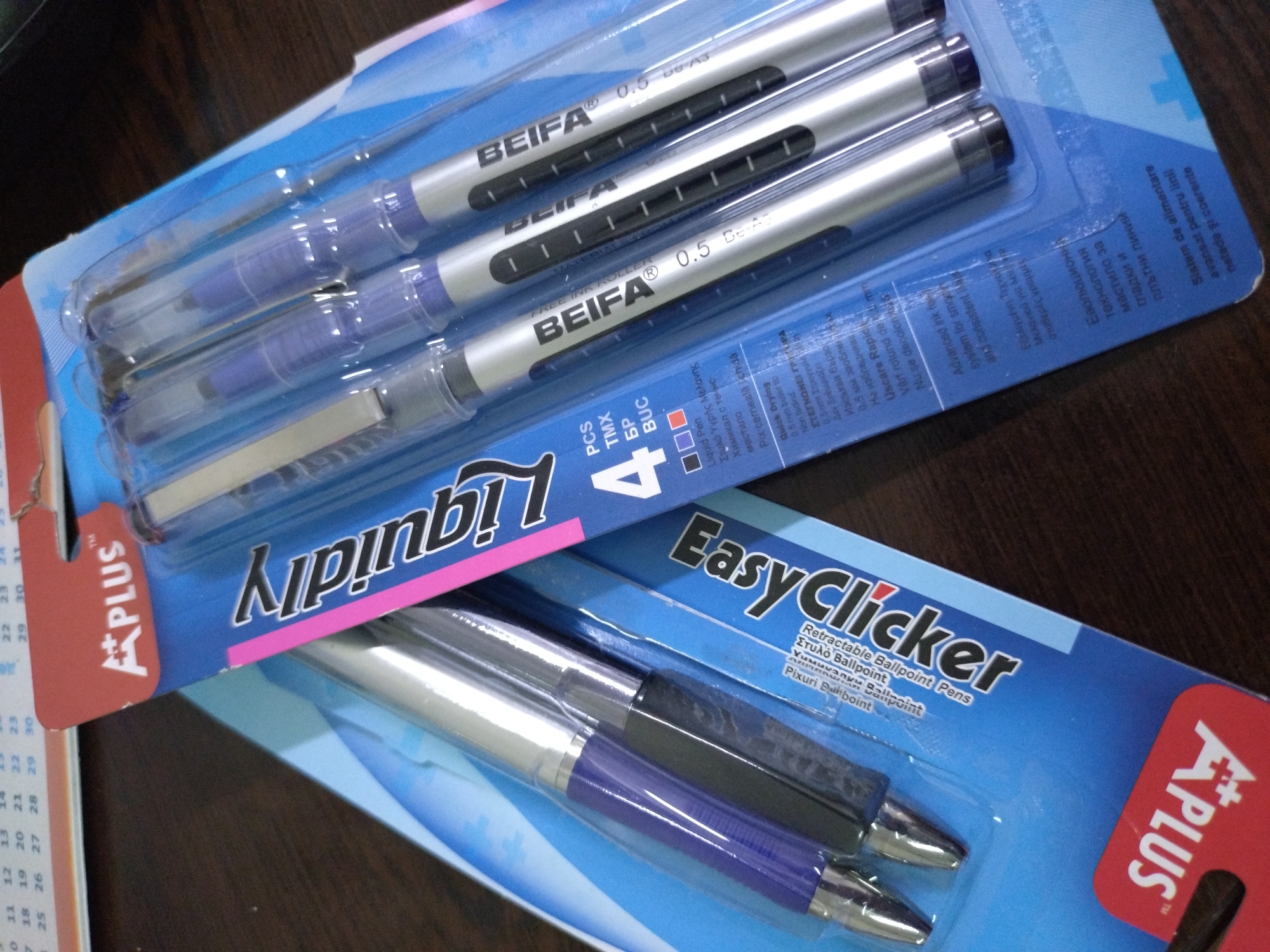 The image shows two packs of pens stacked on top of each other on a wooden surface. The top pack contains four "BEIFA 0.5" free ink roller pens in silver barrels with blue accents. The packaging is blue with "Liquidity" printed prominently. The bottom pack is a pack of "Easy Clicker" retractable ballpoint pens with silver and blue barrels. The packaging is also blue and features the "A PLUS" logo. Part of a calendar with the "A PLUS" logo is visible on the left side of the image.