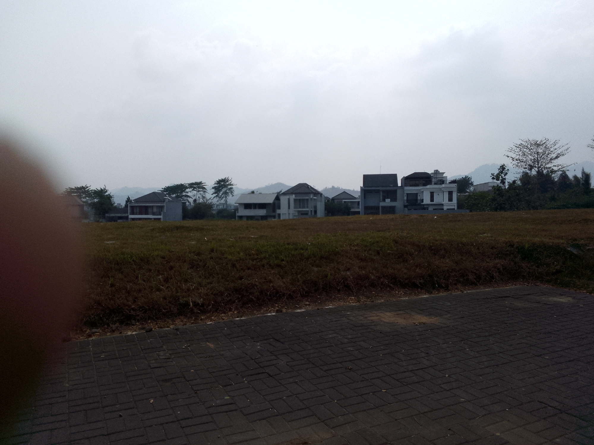 A wide shot captures a suburban residential development with modern architecture under an overcast sky. In the foreground, a dark gray brick-paved path with a subtle diagonal pattern leads the eye towards a brown, dry, grassy field. This field separates the paved area from a row of houses situated on a slight incline in the middle ground. The houses are varied in design, featuring white and gray exteriors, flat roofs, and large windows. Behind the houses, a line of trees, some sparse and bare-branched, with a few larger, leafy trees scattered throughout, partially obscures rolling, green hills in the background. The overall impression is one of a newly developed residential area in a hilly, possibly semi-rural, setting. The sky is a uniform light gray, suggesting a cloudy or foggy day. The scene is static and peaceful, with no visible people or activity.