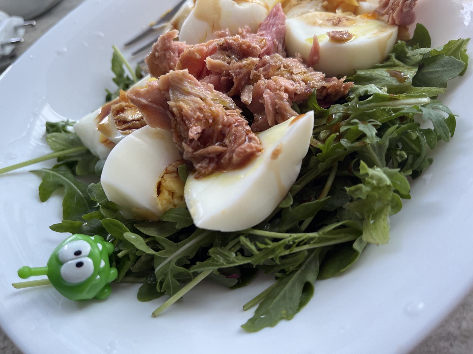 An overhead, close-up shot shows a white salad bowl filled with a salad of arugula, tuna, and hard-boiled eggs. A small green plastic toy, resembling the character Om Nom from the game Cut the Rope, is stuck into the salad near the top left. The salad is dressed and appears to have a light sheen from the dressing. The background is blurred and shows a hint of a fork and another white surface, possibly a table or another plate. The image is brightly lit, suggesting it was taken indoors with natural light. There are no people visible in the image, and the location is unknown.