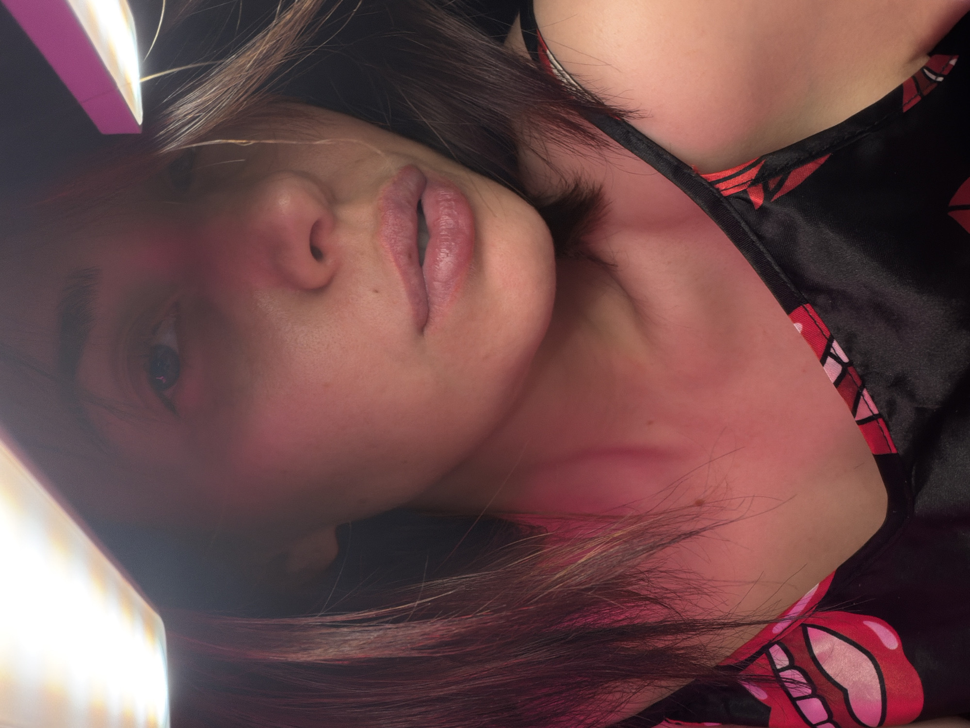 In a close-up, low-angle shot, a young woman's face is illuminated by bright pink and white lights from above, creating dramatic shadows. She has medium-length, dark brown hair that frames her face, with some strands falling across her cheek. Her eyes are partially visible, looking slightly upwards. Her lips are slightly parted, revealing a subtle sheen. She is wearing a black camisole with a pattern of red and pink lips. The background is dark and indistinct, with the lights creating a halo effect. The scene appears to be indoors, possibly in a bedroom or a studio setting, with no discernible urban or natural elements. The overall lighting suggests a deliberate setup for photography or video.