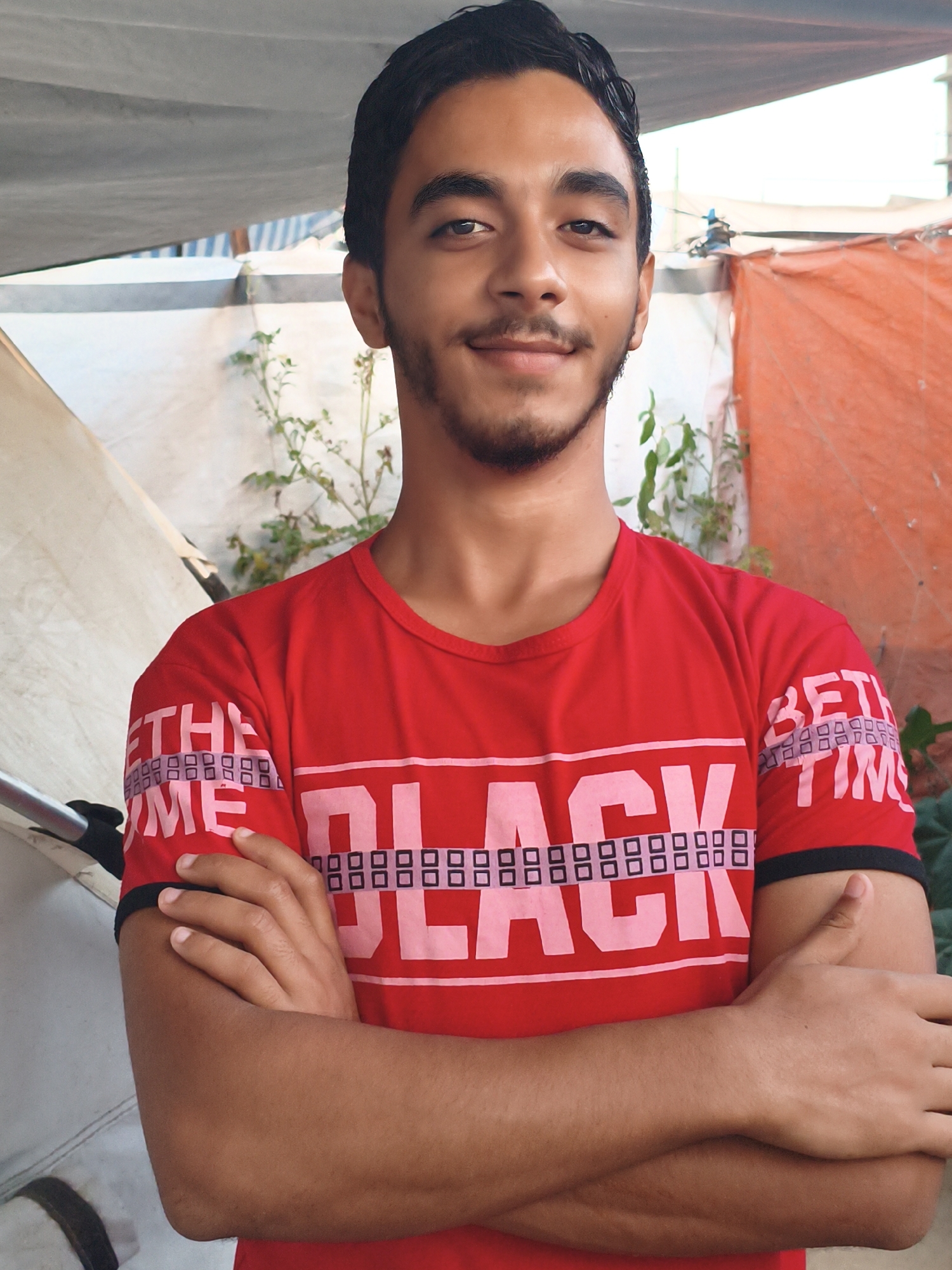 A young man with dark hair and a light beard is pictured from the chest up, facing the camera with his arms crossed. He is wearing a red t-shirt with large pink letters on it that spell out "BLACK" in the center, and "THE TIME" on the sleeves in a smaller font. He appears to be standing outdoors, with a light-colored awning or tarpaulin overhead and some greenery and orange fabric in the background. The scene is likely in the city of Gaza, though the exact location is unknown.