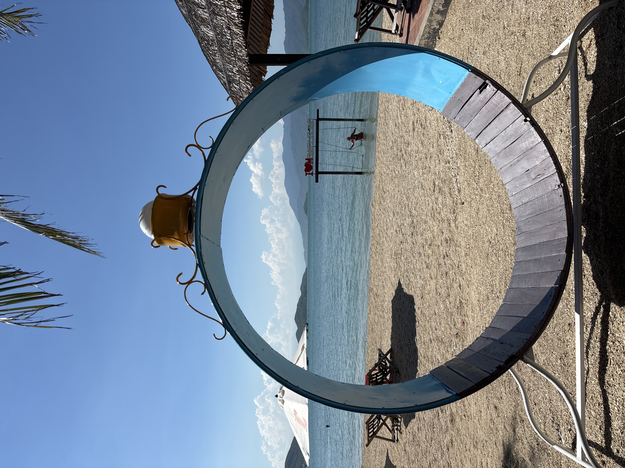 A beach scene in Vĩnh Lương, Vietnam, features a large, circular, blue-and-wood sculpture in the foreground. The sculpture is supported by a white metal frame. Beyond the sculpture, a sandy beach stretches to the water, with two empty lounge chairs visible. In the distance, a person is on a swing set in the shallow water, with a sign that reads "I LOVE MONKEY ISLAND." The sea meets a mountainous horizon under a clear blue sky with scattered clouds. Palm fronds hang from the top of the frame.