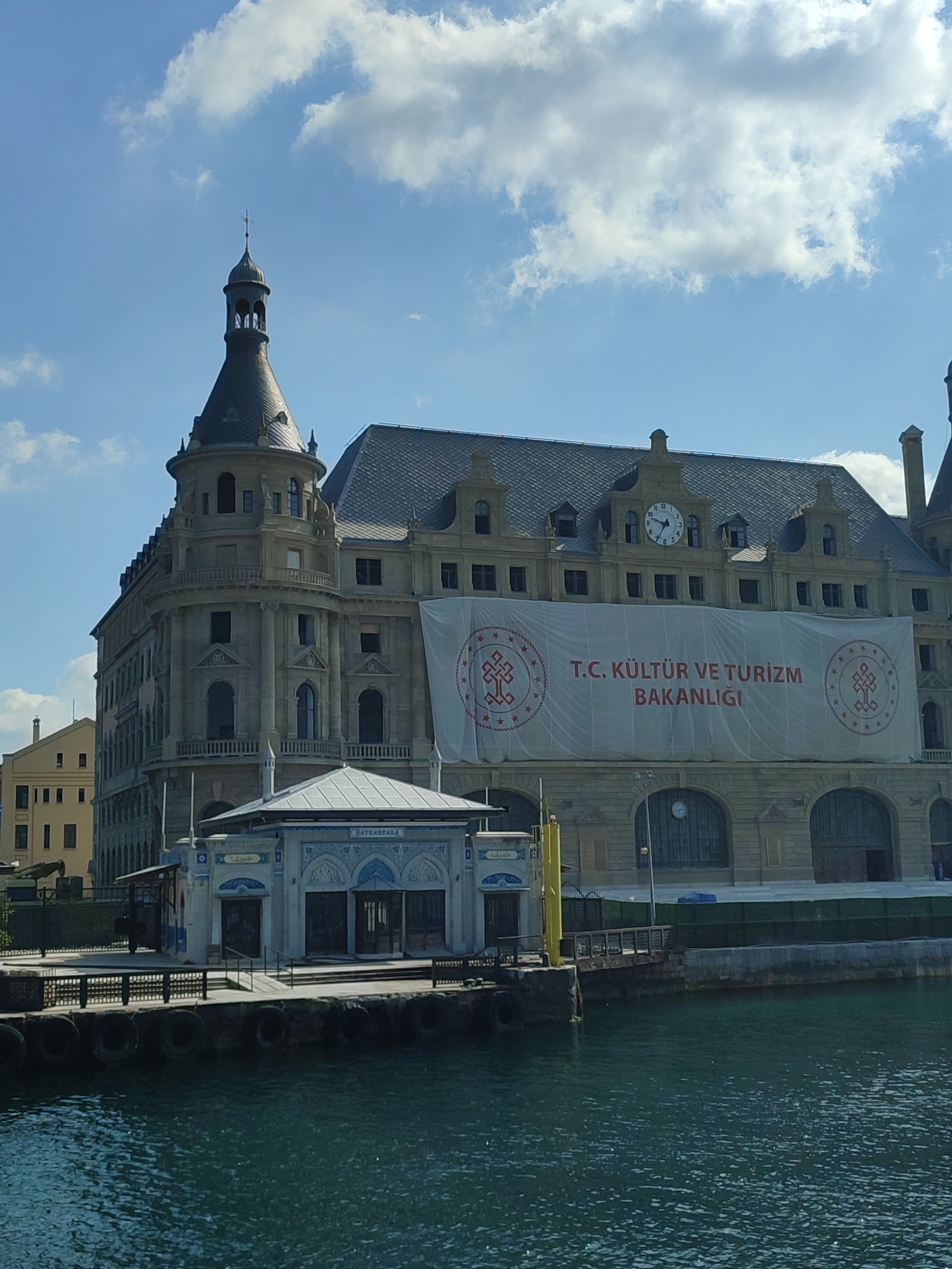 This image captures a portion of the historic Haydarpaşa Train Station in Kadıköy, Turkey, on a bright, partly cloudy day. The station, a prominent landmark on the Asian side of Istanbul, is characterized by its imposing Beaux-Arts architecture. A large white banner with red text and insignia hangs across the facade, displaying "T.C. KÜLTÜR VE TURİZM BAKANLIĞI" (Republic of Turkey Ministry of Culture and Tourism).
In the foreground, the dark, rippling water of the Bosphorus is visible, with the weathered concrete embankment of the quay. A small, ornate building with blue and white tile accents, likely a ticket booth or waiting area, is situated on the quay. Fenders made of tires are attached to the embankment to cushion boats.
The train station itself features a distinctive clock tower, visible in the upper left of the image, topped with a cross-like finial. The roof is steeply pitched and covered in dark tiles. Several arched windows and balconies punctuate the building's facade. To the left, a smaller, lighter-colored building stands behind the train station, and beyond that, a yellow building with many windows. A yellow crane-like structure is partially visible on the left side of the image, near the water's edge. The sky is a vibrant blue, with scattered, fluffy white clouds. The scene conveys a sense of historical significance and bustling maritime activity.