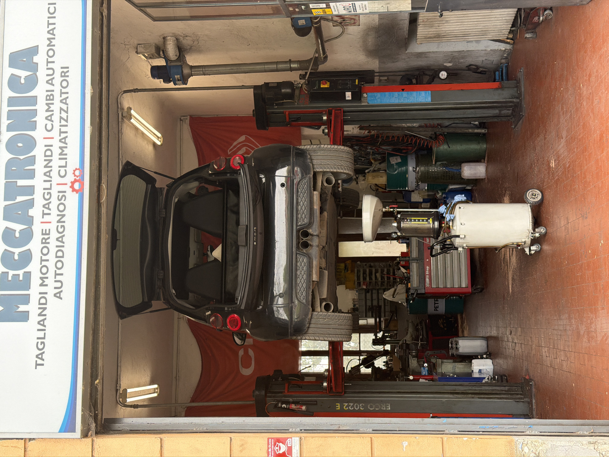 The image captures the interior of an auto repair shop. A black vehicle is elevated on a hydraulic lift. Tools and equipment are arrayed around the car. A prominent sign reading "MECCATRONICA," with Italian text indicating services like "TAGLIANDI MOTORE," "CAMBI AUTOMATICI," and "CLIMATIZZATORI" is on the left. A red Citroen sign on the lift and a red sticker reading, “Guardia Giurata Pronto Intervento 24h”, which translates to Security Guard Urgent Response 24h, suggest that the location may be in Italy. Based on the Italian text, the location is likely in the city of Milan, Italy.
