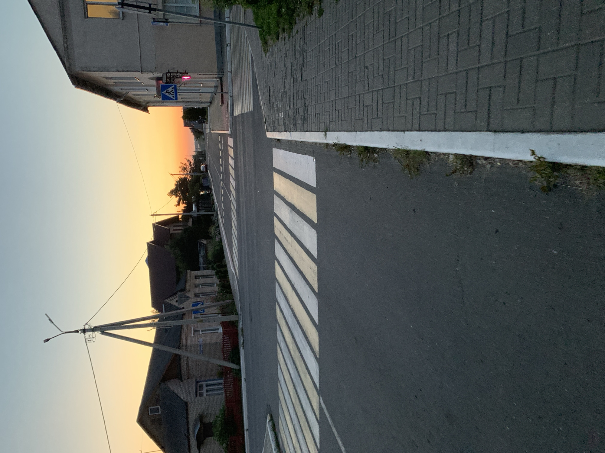 The image depicts a street scene at dusk in Kapyĺ, Belarus. A crosswalk with alternating white and yellow stripes is prominently featured in the foreground. To the right, a paved sidewalk with gray brickwork runs along the street. Adjacent to the sidewalk is a building with a light-colored facade. A street lamp on a concrete pole stands to the left of the street, with wires extending across the sky. Houses with brick walls and dark roofs are visible on the left side of the street. The sky is a gradient of pale blue at the top to orange and yellow near the horizon, indicating the setting sun. A traffic light with a red signal is visible near the building on the right. The road surface is asphalt. The overall impression is one of a quiet, residential street at the end of the day.
