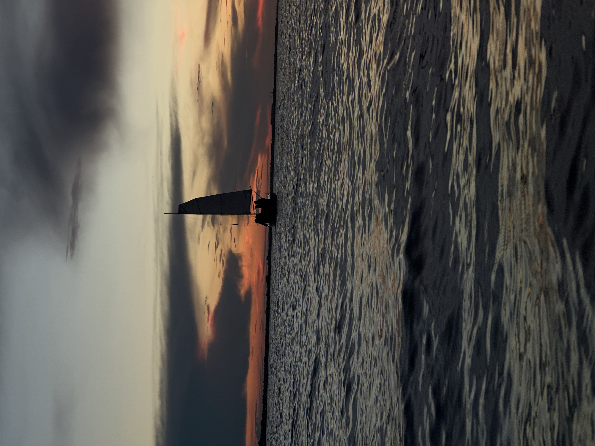 This image depicts a sailboat on a body of water during a sunset. The sky is filled with dark clouds tinged with orange and pink hues, indicating the sun is low on the horizon. The water is choppy, with visible ripples. A dark silhouette of the sailboat, with a single sail and several figures on board, is positioned slightly to the left of the center of the frame. The distant horizon shows a faint line of land. The overall atmosphere is one of tranquility and the end of the day.
There is no indication of the city of Kokshetau, Kazakhstan, in this image.