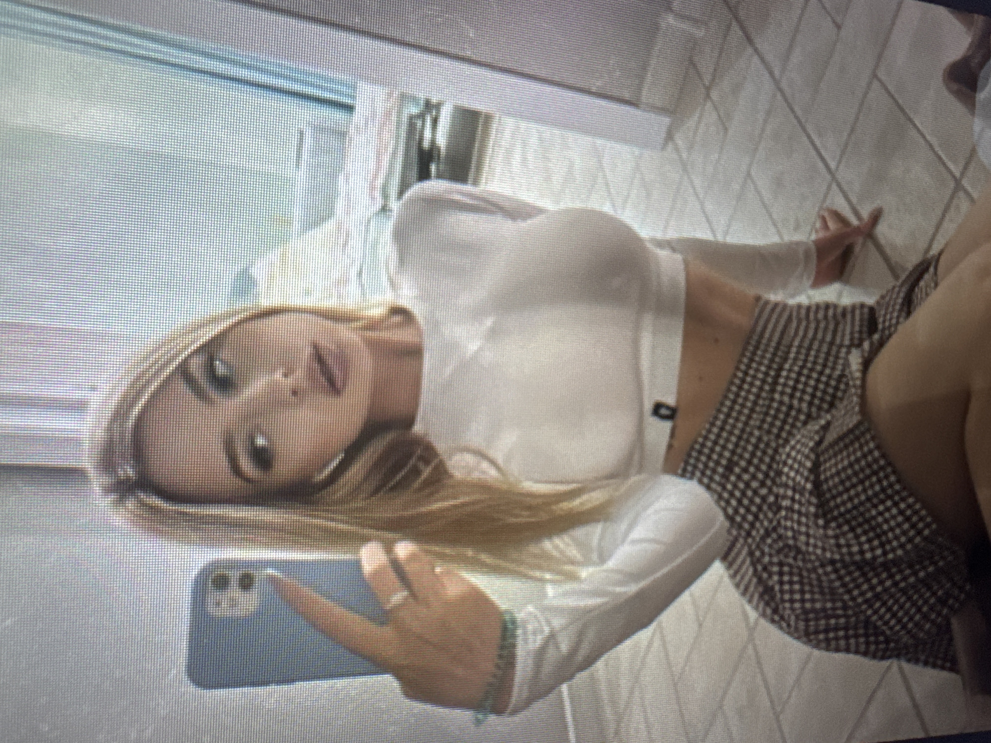 A pixelated, rotated image depicts a young woman with long blonde hair, appearing to take a selfie in an indoor setting, possibly in Varna, Bulgaria. She is positioned on her side or leaning back, holding a light blue smartphone in her right hand towards herself. She wears a form-fitting white long-sleeved crop top that exposes her midriff, paired with a brown and white gingham or plaid mini-skirt. A ring is visible on one of her fingers and a green beaded bracelet on her wrist. The woman looks directly at the phone's camera with a posed expression. The background includes light-colored tiled flooring laid in a diagonal pattern, and a white bed or mattress with a quilted pattern is partially visible. No discernible text is present.