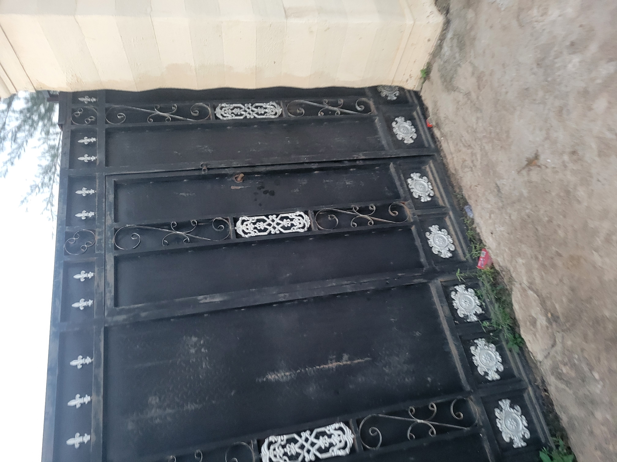 The image shows a black wrought iron gate in front of a light-colored stone wall. The gate has ornate decorative elements, including fleur-de-lis motifs at the top and circular patterns and scrollwork on the panels. The gate appears to be partially open or slightly ajar. The ground in front of the gate is a concrete or stone surface, with some dirt and small plants growing along the base of the gate. A few small pieces of litter are visible on the ground near the gate. The scene is outdoors, and the lighting suggests it is daytime, possibly overcast. There are no people visible in the image.