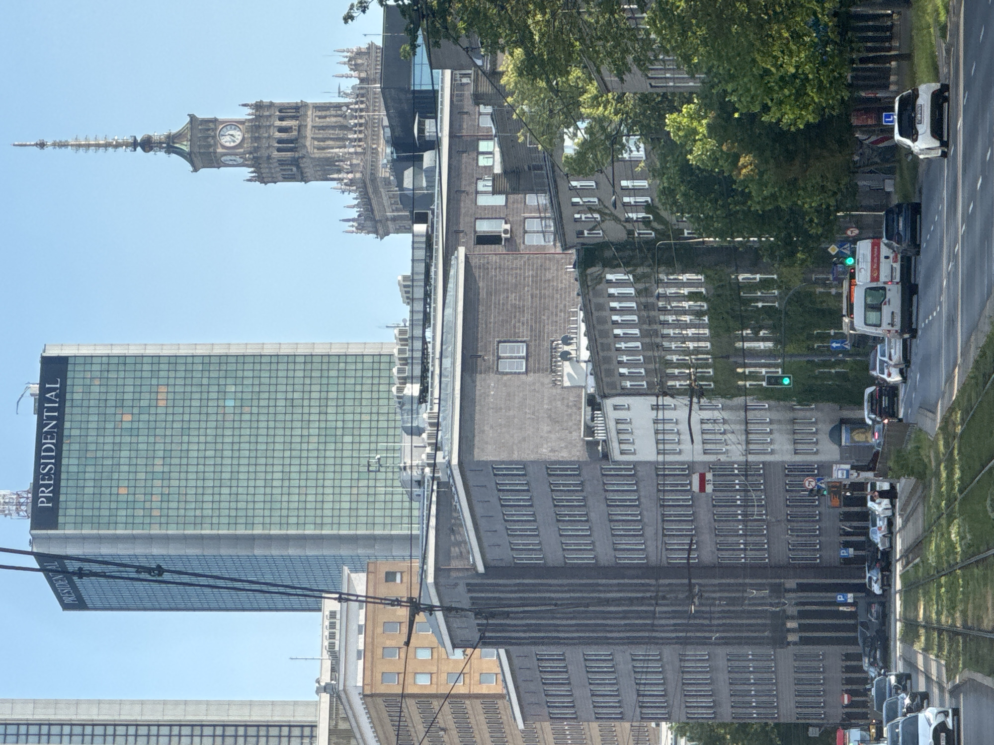 This is a daytime, medium-distance, eye-level shot of a street scene in Warsaw, Poland, featuring multiple buildings and a street with traffic. In the background, the Presidential Building, a tall glass skyscraper with a grid facade, is prominently visible, displaying the word "PRESIDENTIAL" at its top. To the right of it, the Palace of Culture and Science, a grand, historic building with a pointed tower and a clock face, rises against a clear blue sky. In the foreground and midground, several lower, dark-colored buildings with numerous windows line the street. A tram track runs through the middle of the street, which is populated by various cars, vans, and a bus. Lush green trees are interspersed throughout the scene, particularly along the right side of the street and between buildings. The overall impression is a blend of modern and historic architecture in a busy urban environment.
